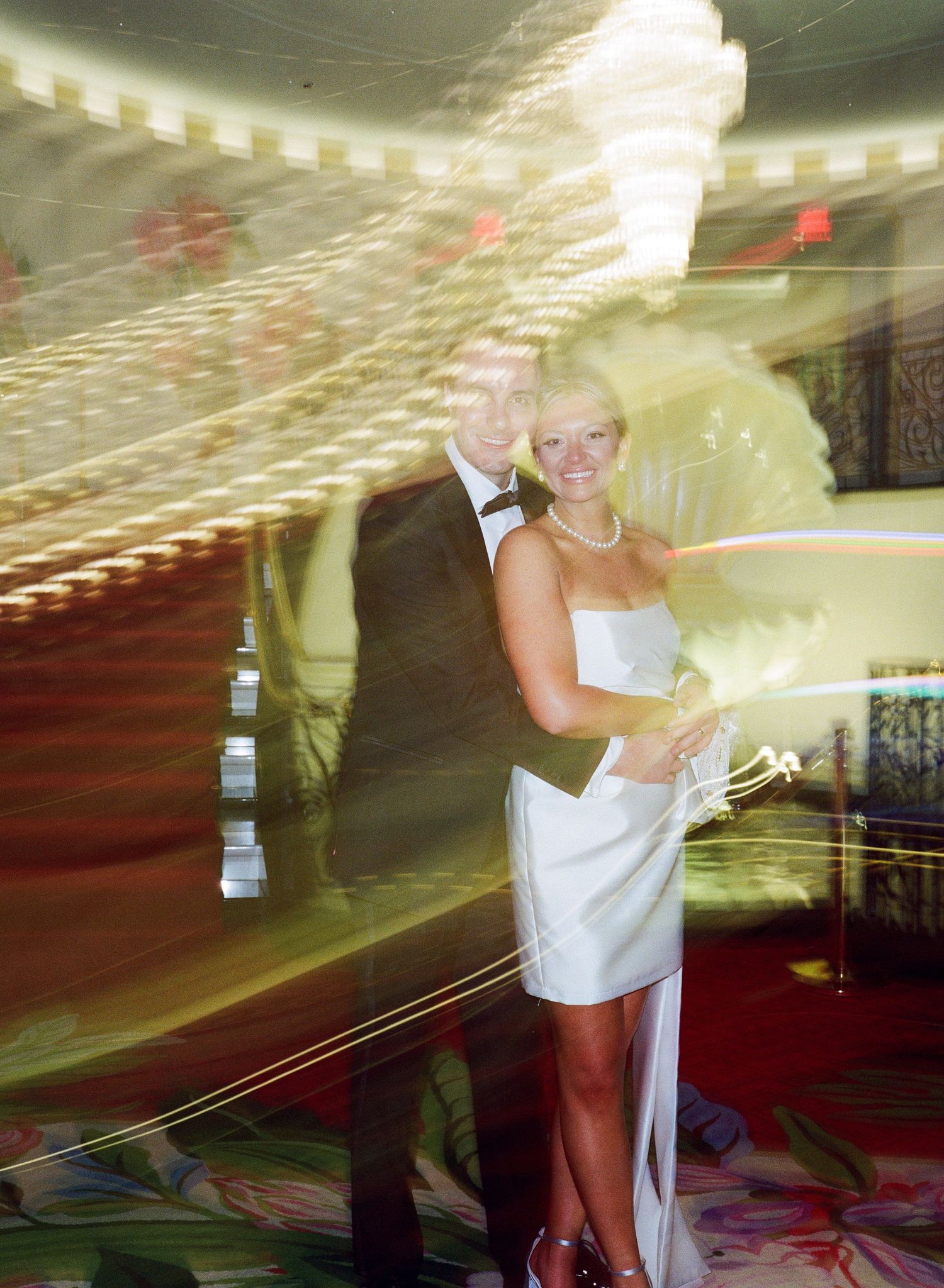 Couple in formal wedding attire posing together, woman in white dress with pearl necklace, man in tuxedo, motion blur effect.