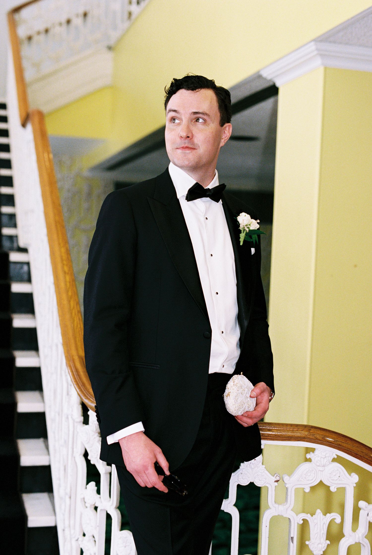 Groom in black tuxedo with boutonniere stands by elegant staircase holding bridal accessories at wedding venue.