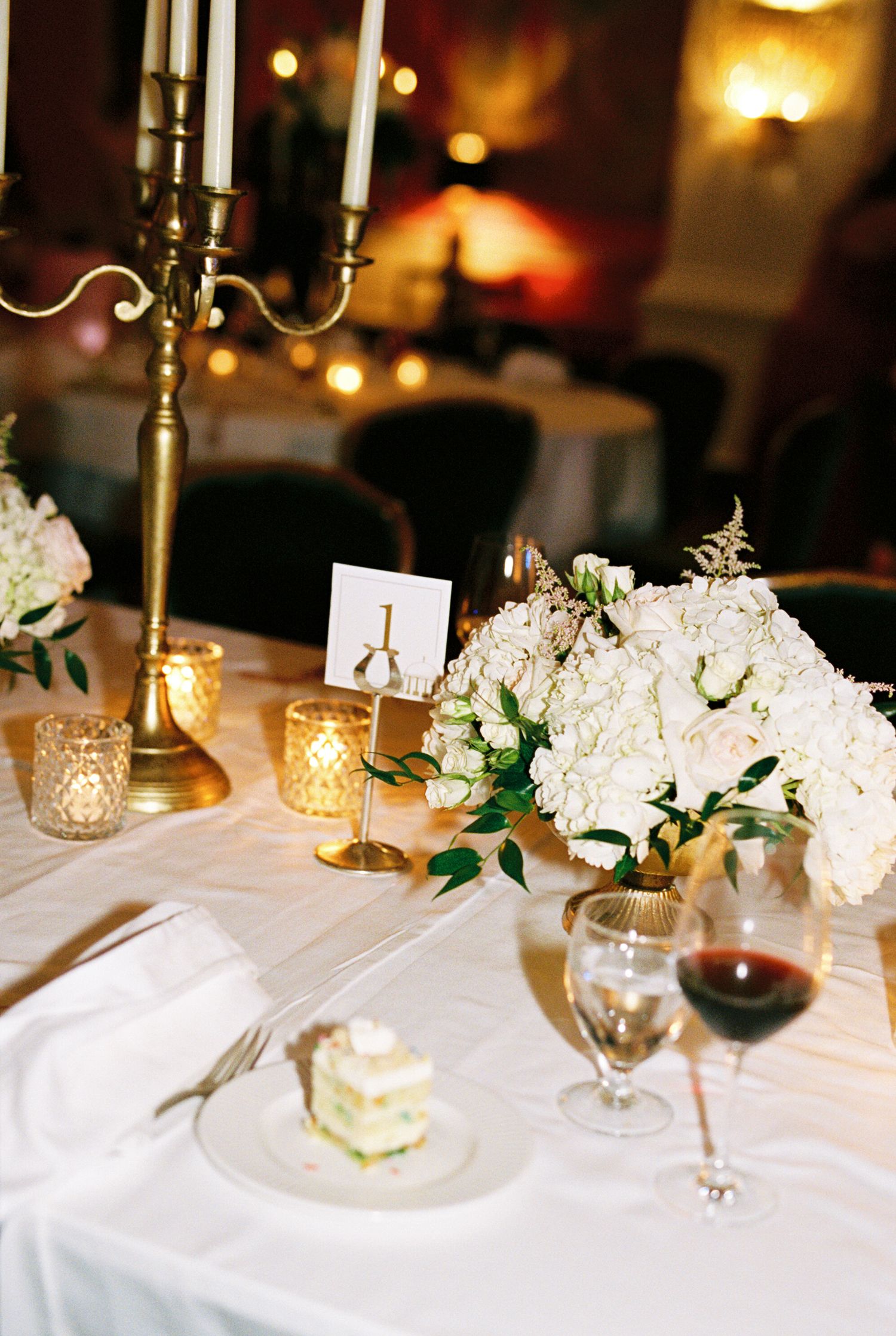 Elegant wedding reception table with white floral centerpiece, gold candelabra, red wine glass, and slice of cake.