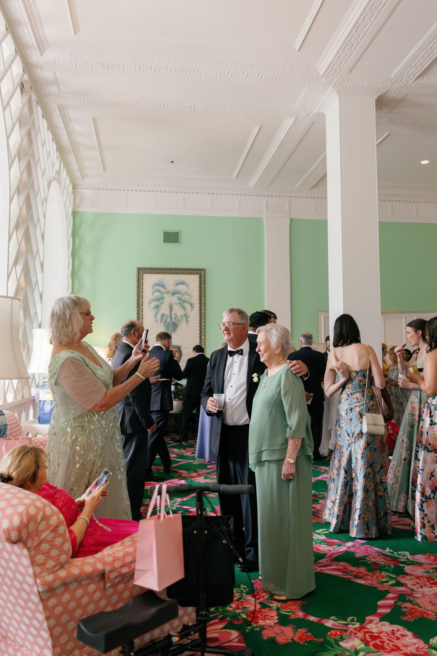 Elegantly dressed guests mingle in a green-walled ballroom during a formal event or wedding reception.