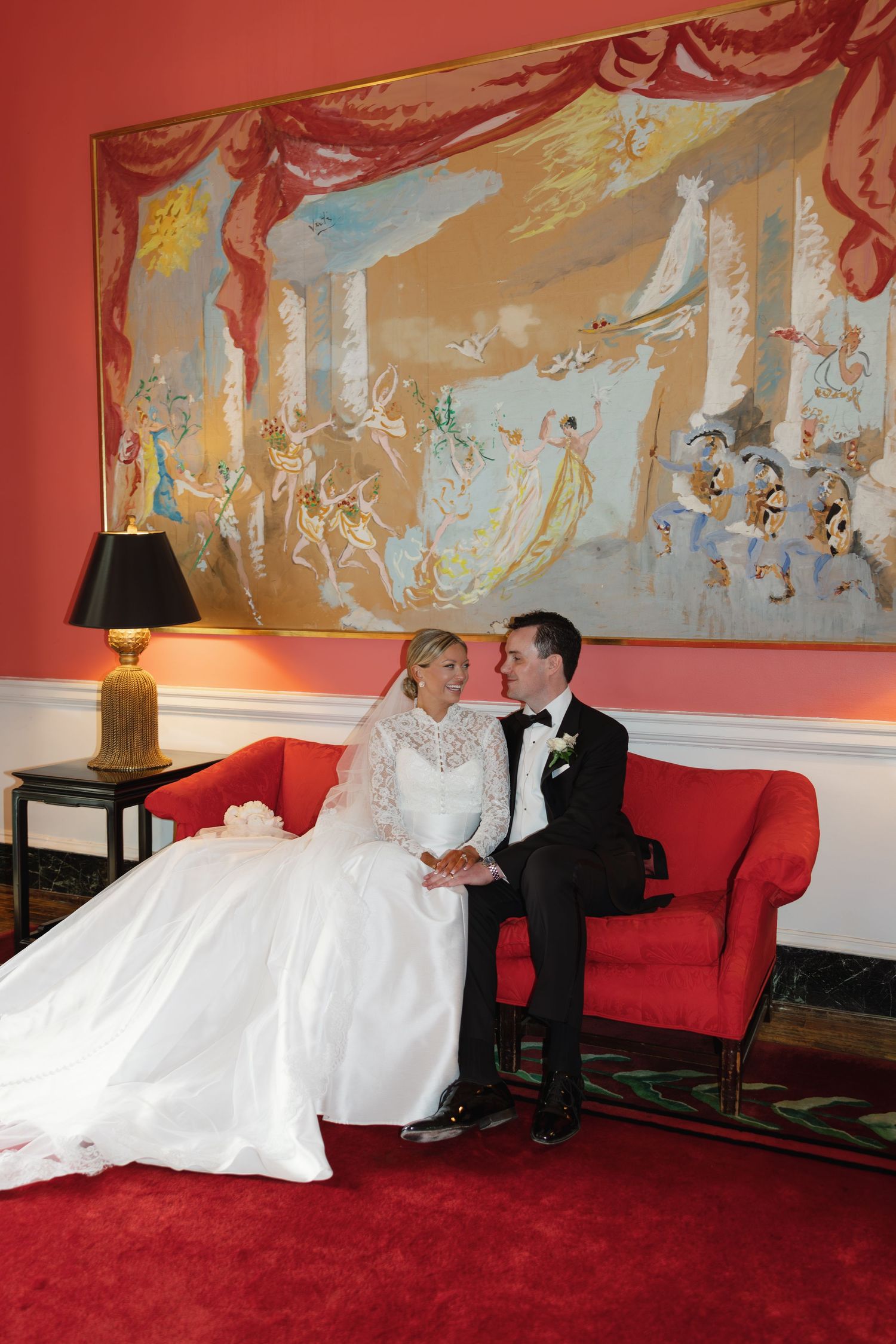Bride and groom sit on red sofa holding hands before colorful mural in elegant red-walled wedding venue.