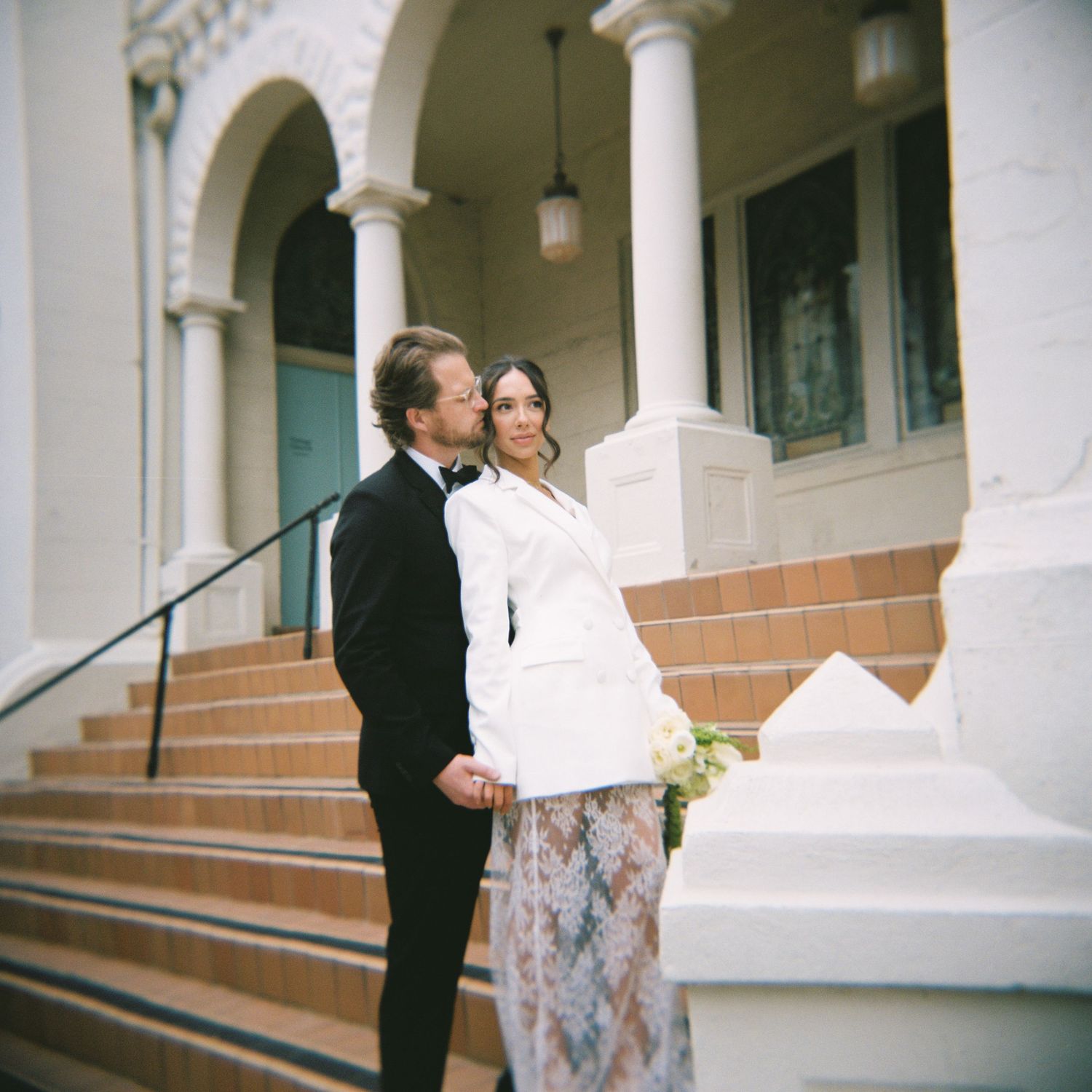 Elegant wedding couple posing on brick steps outside a white columned building, bride holding a bouquet in a white blazer.