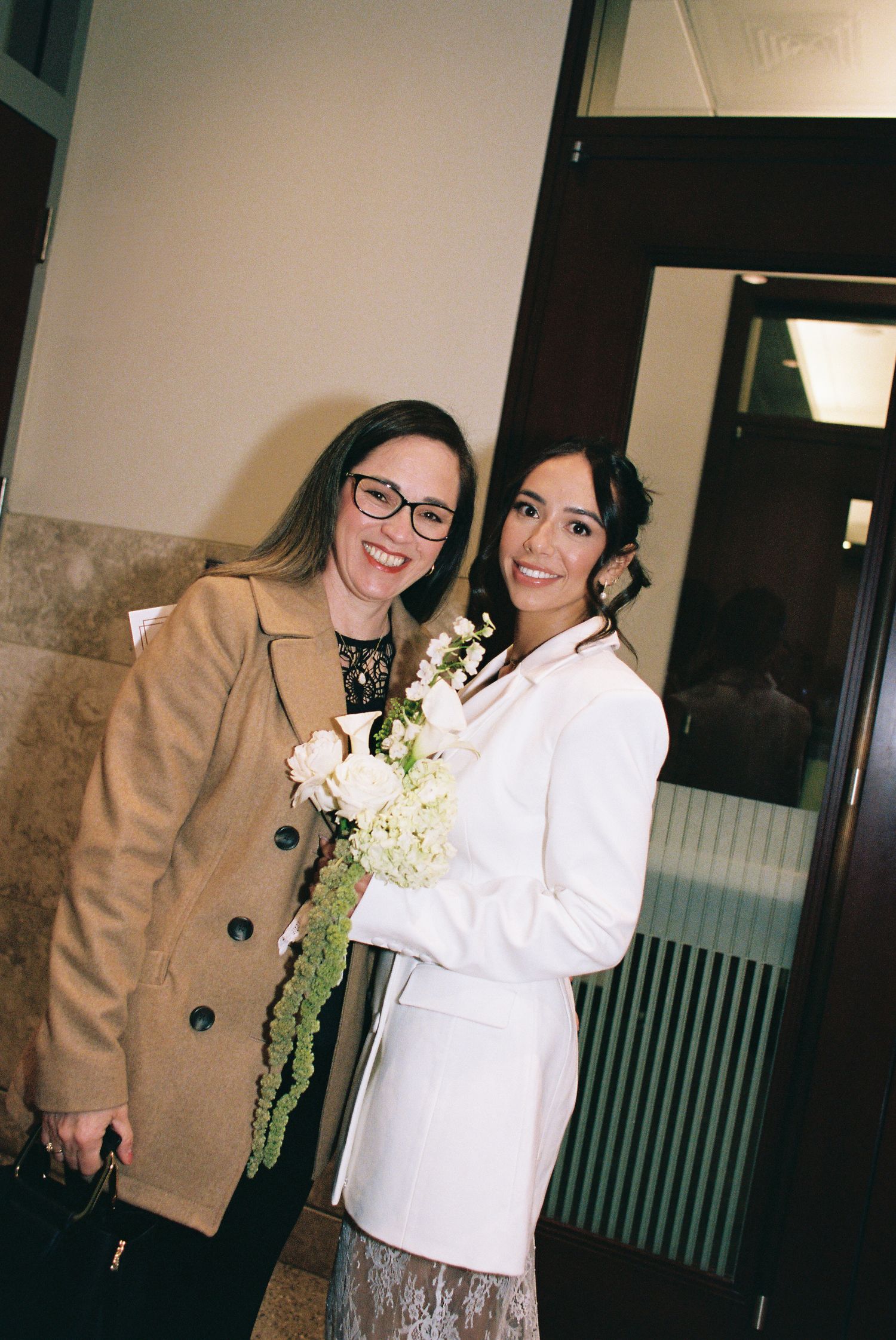 Two women posing together, one in a white blazer holding a bouquet of white flowers, likely at a wedding venue.