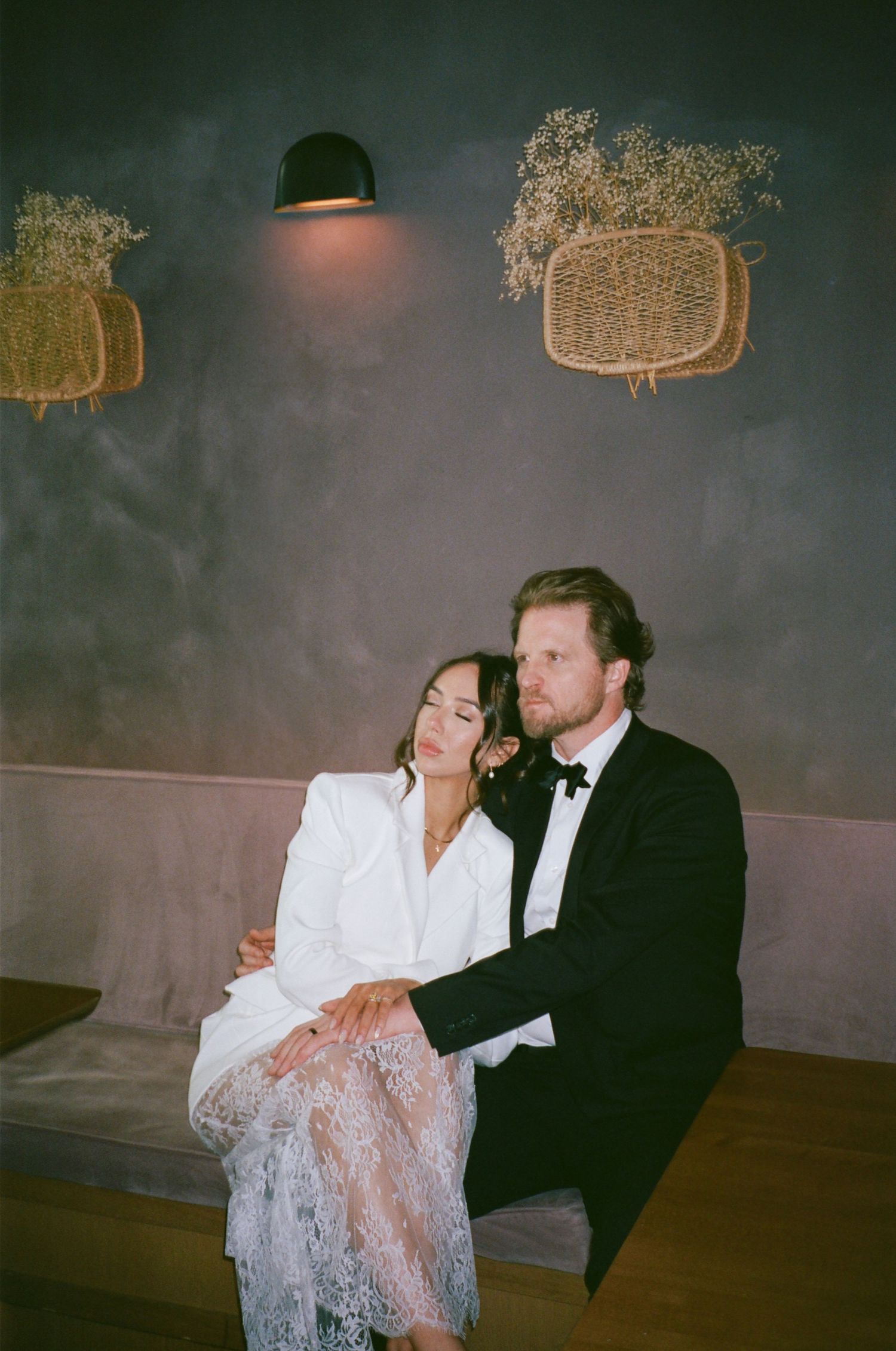 Elegant couple in wedding attire sitting together on a bench, woman in white lace dress, man in black tuxedo.