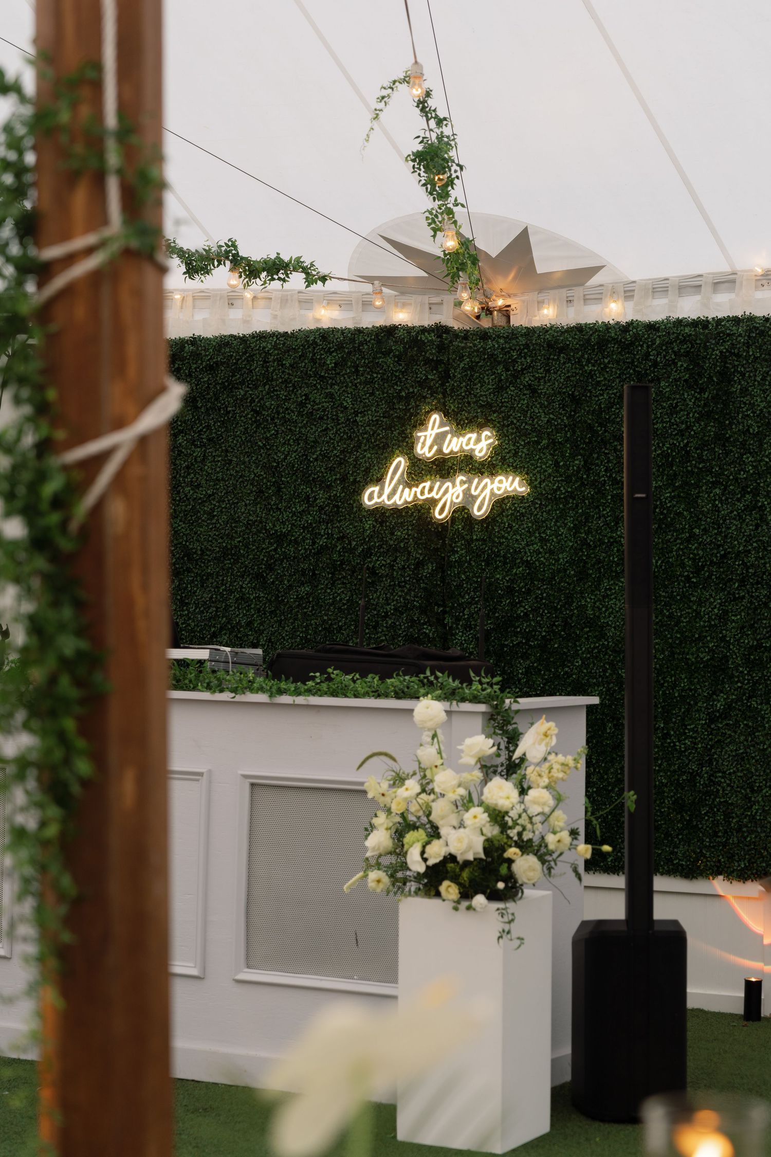 Elegant wedding venue decor featuring illuminated neon signage against a lush green hedge wall with white floral arrangements.