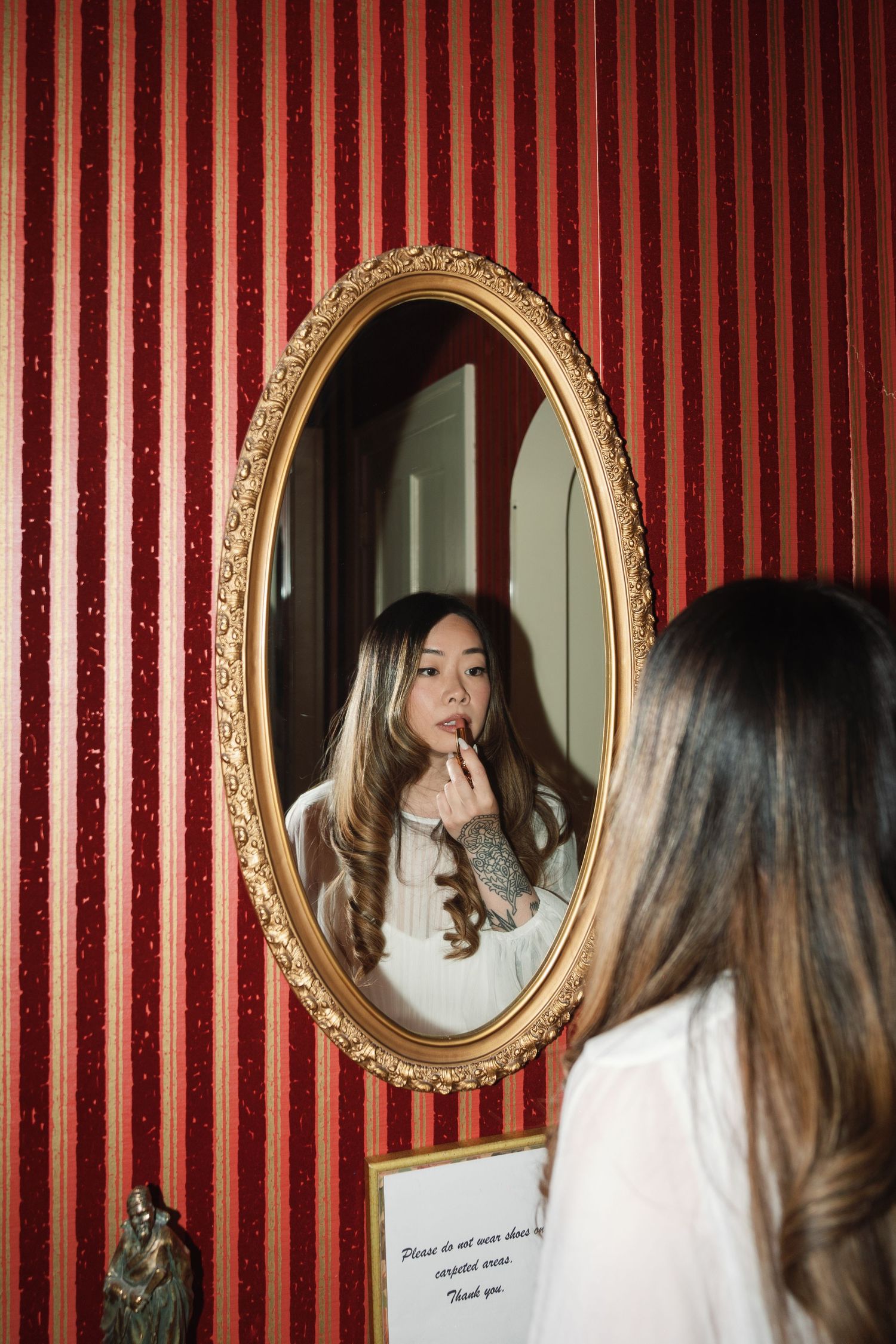 A reflection in an ornate gold-framed oval mirror mounted on red striped wallpaper shows long wavy hair and thoughtful pose.