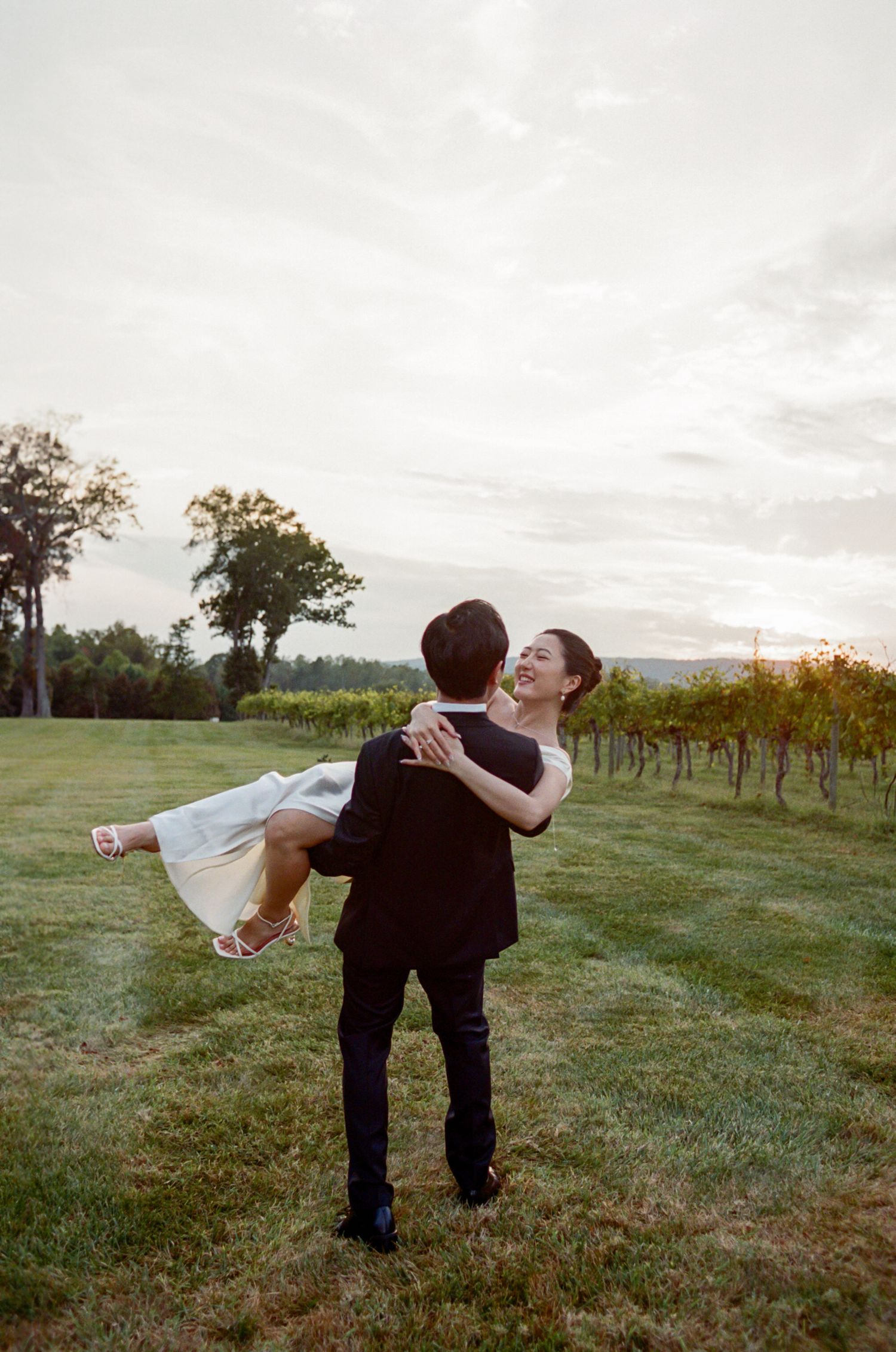 Romantic couple embraces in a sunset vineyard setting as one carries the other in their arms on a grassy hill.