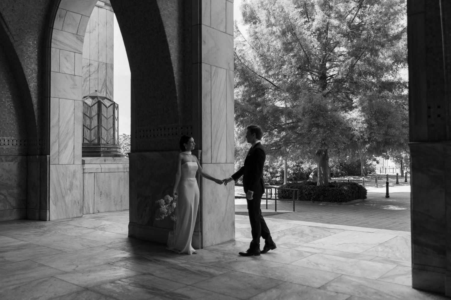 Black and white photo of a bride and groom holding hands beneath stone archways, with trees and an ornate window in the background.