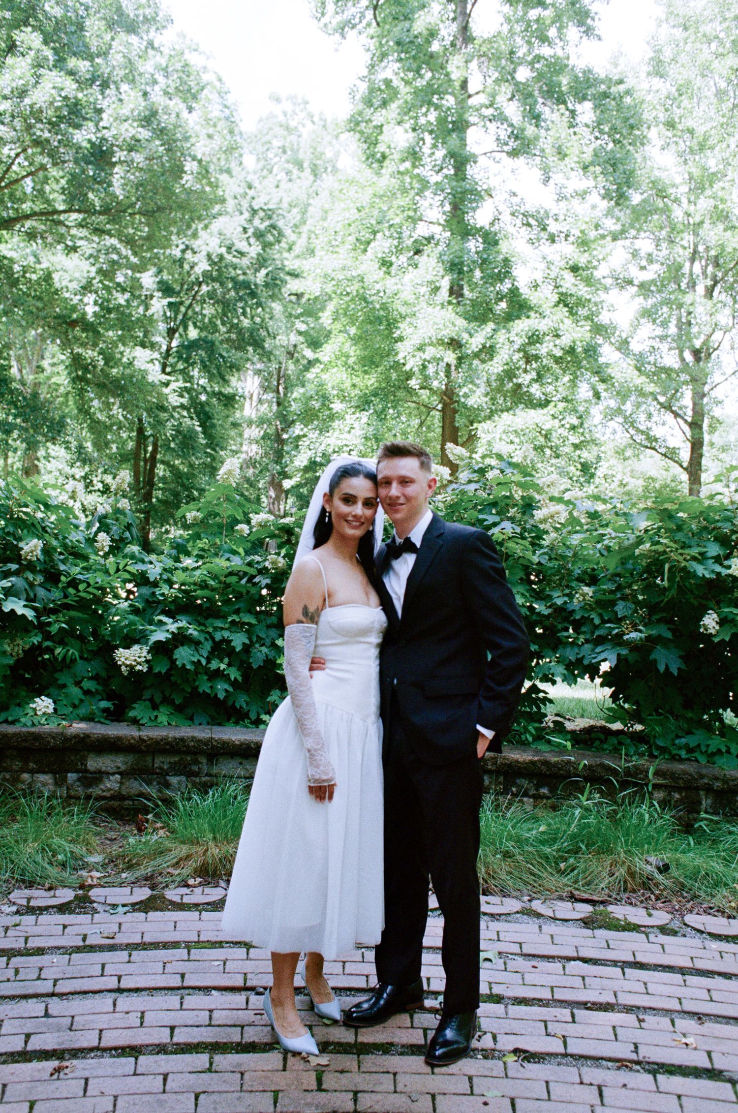 Bride in a short white dress with veil and groom in a black tuxedo and bow tie embrace on a brick patio surrounded by lush green trees.