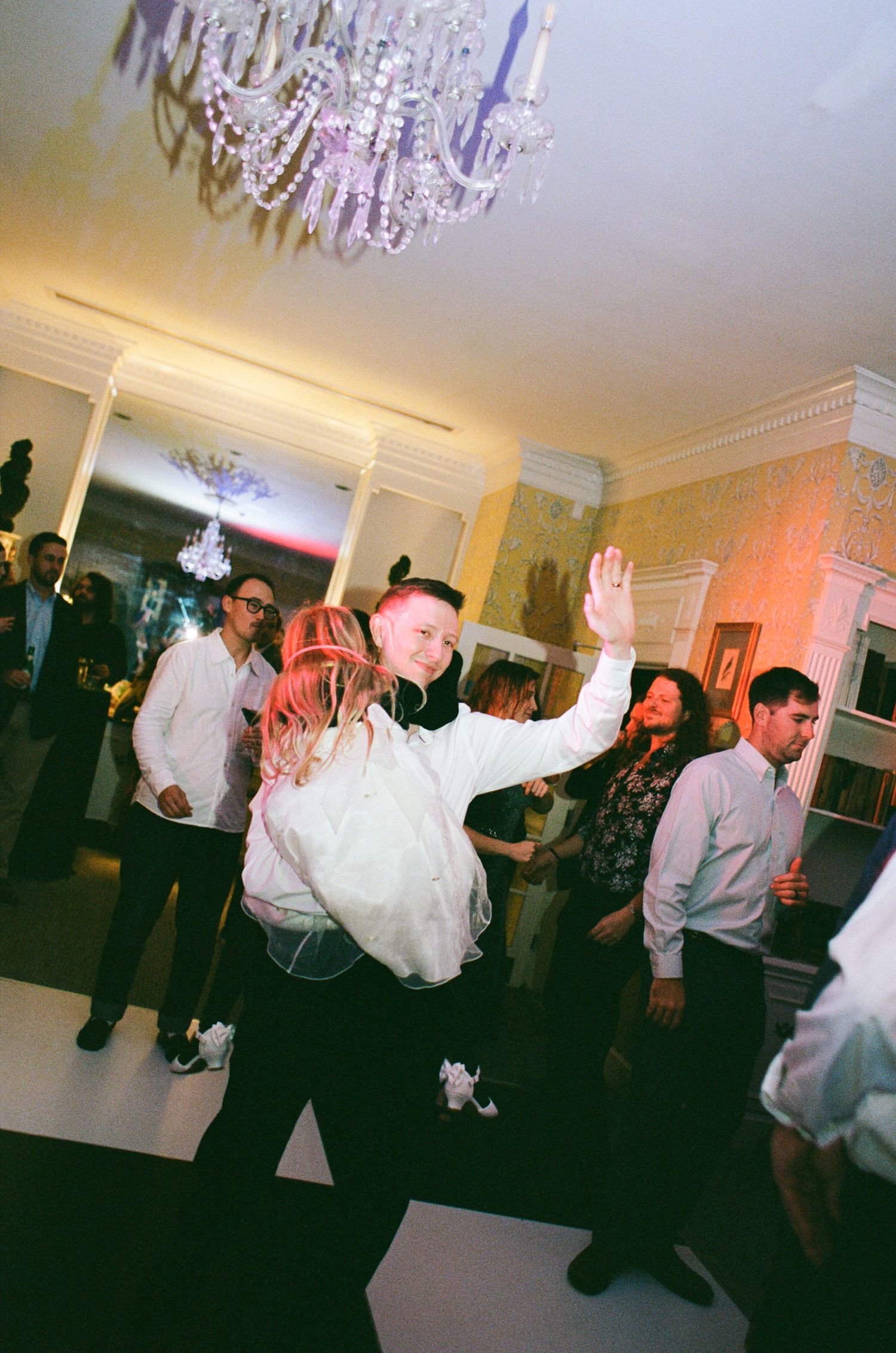 Wedding guests dance and celebrate on a dance floor beneath a sparkling crystal chandelier in an elegant ballroom.