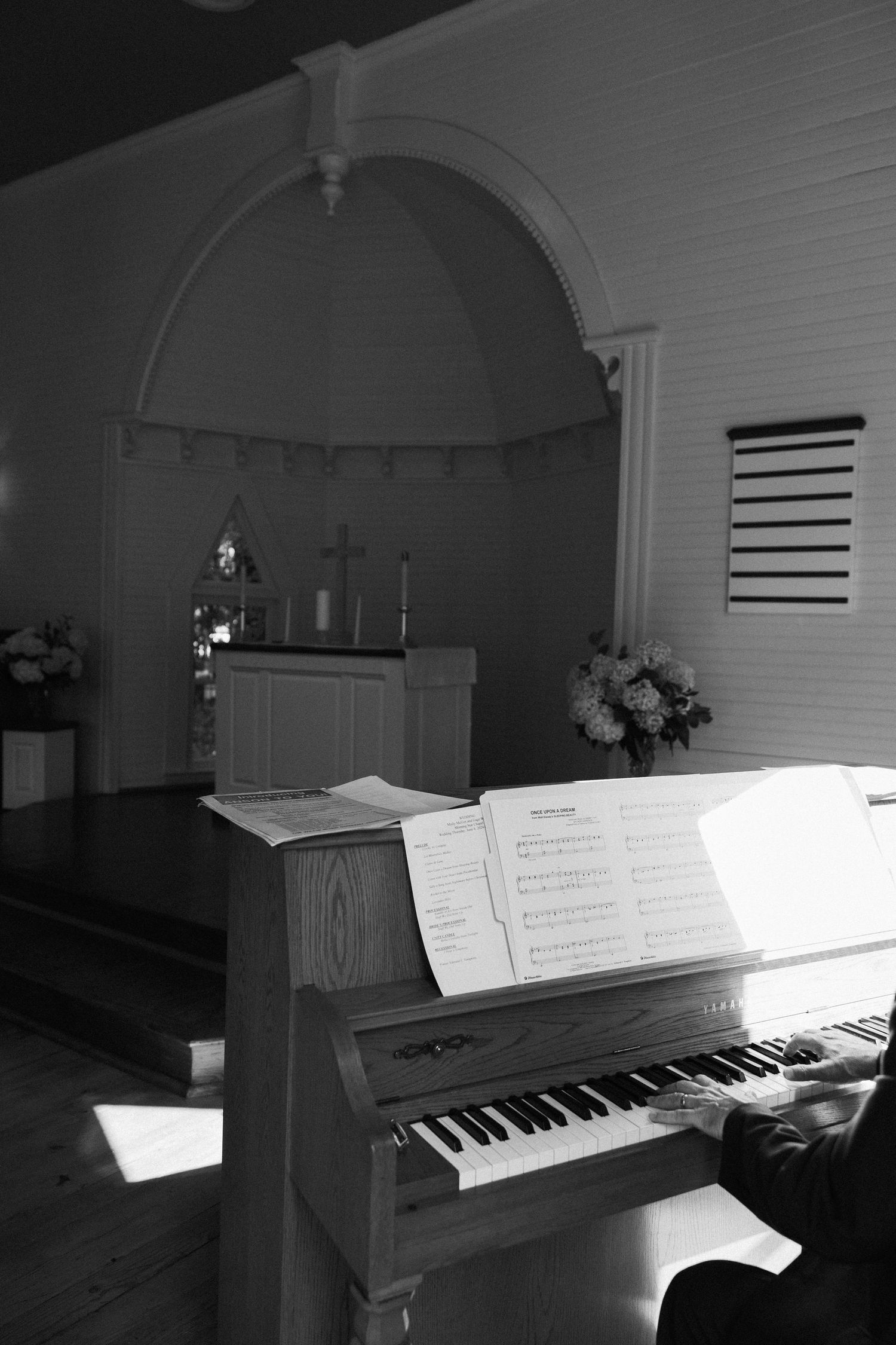 Black and white photo of a person playing an upright piano with sheet music in a small chapel with an altar, cross and flowers.