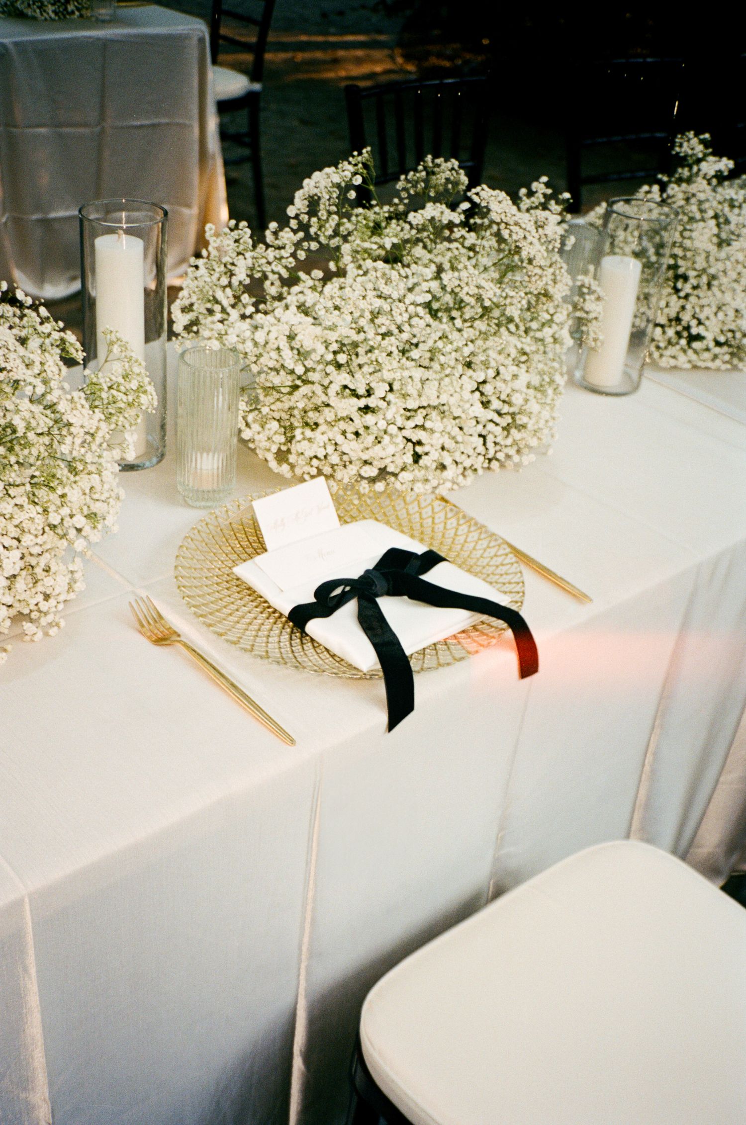 Wedding table setting with gold charger plate, napkin tied with black ribbon, gold flatware, baby's breath centerpieces and white candles.
