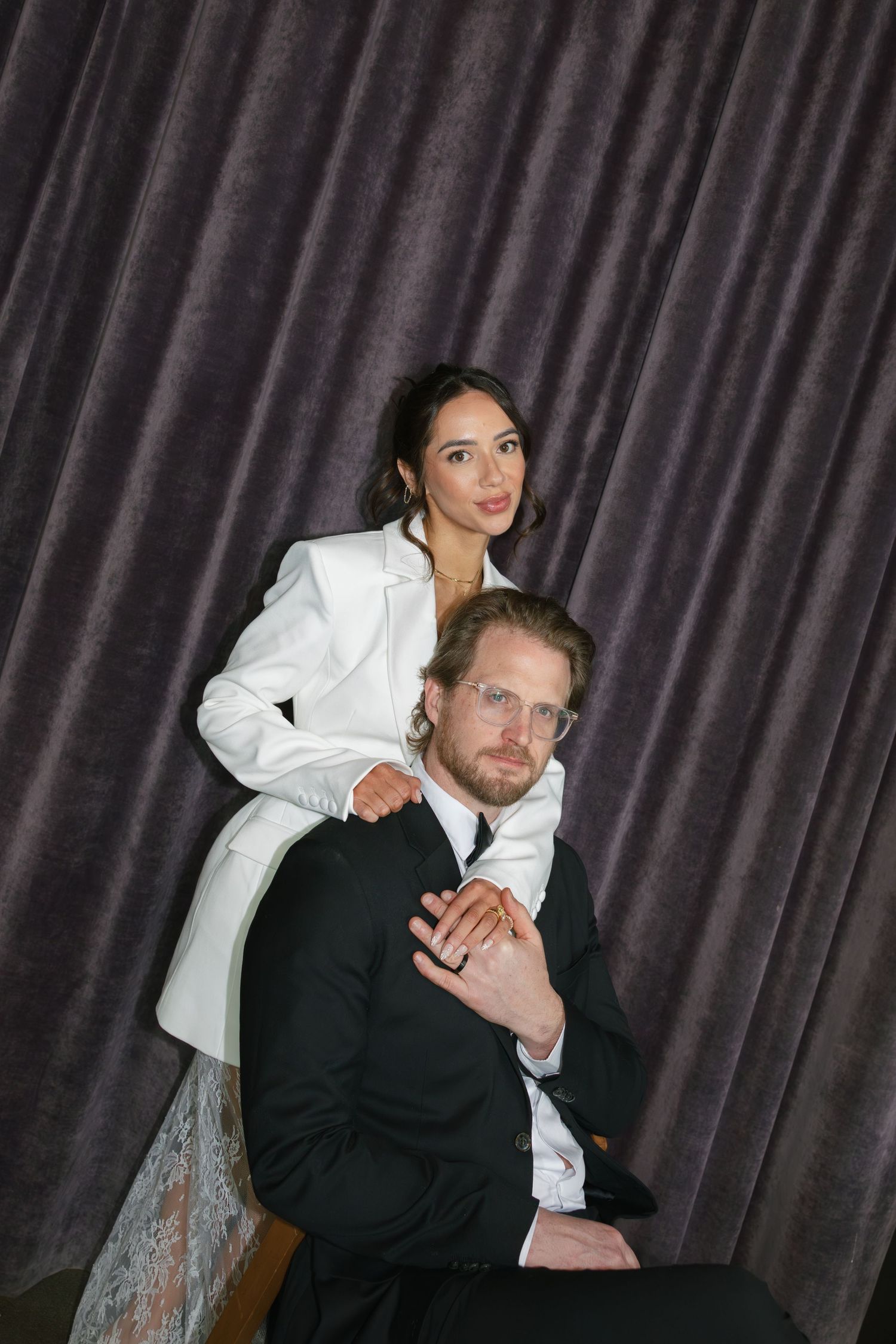 Elegant couple posing against gray velvet curtain, woman in white blazer stands behind seated man in black suit with glasses.