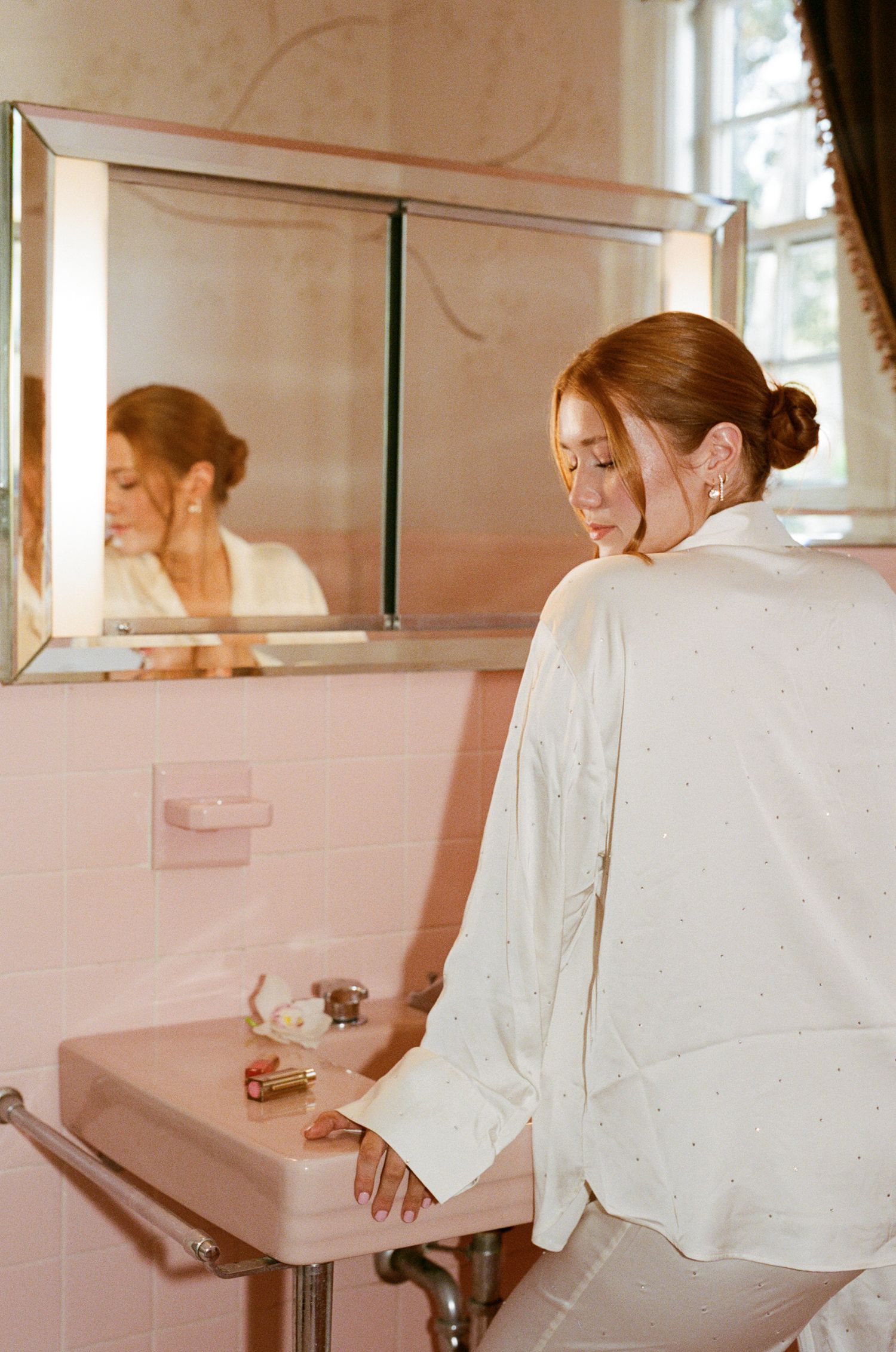 Woman in white satin blouse at pink vintage bathroom sink, reflected in mirror.
