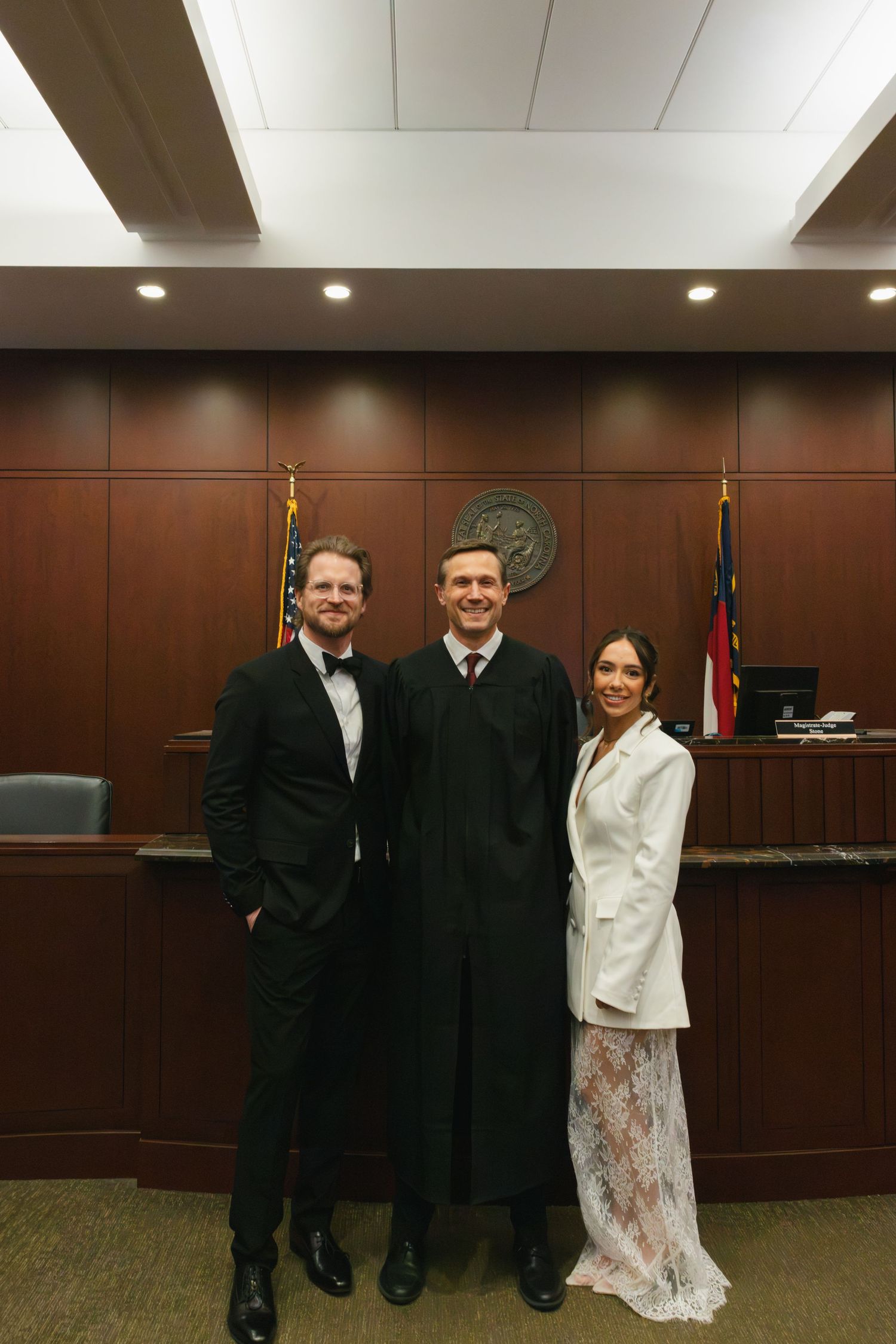 Couple in formal attire posing with a judge in black robes inside a courtroom for a civil wedding ceremony.