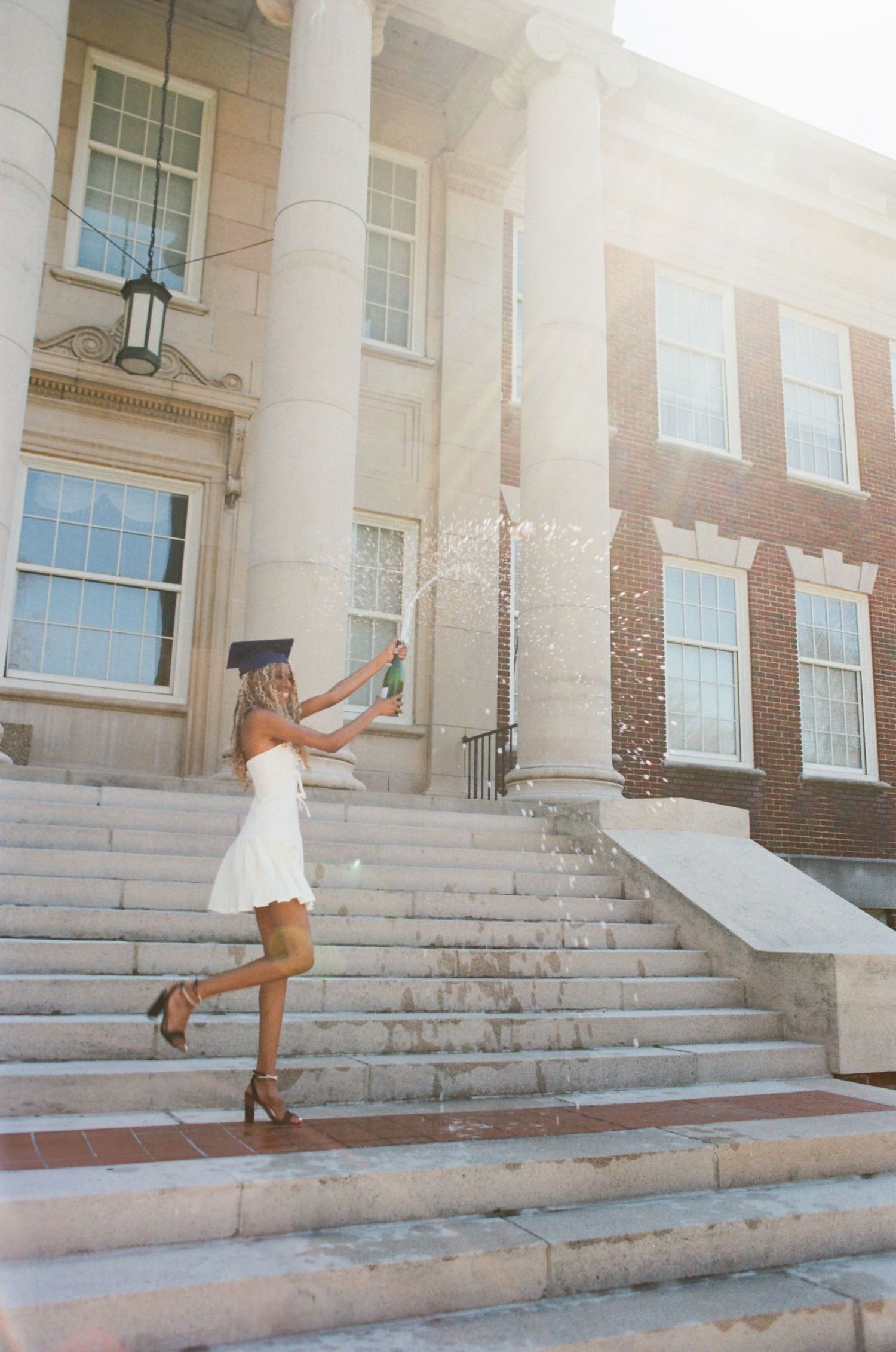 Graduate celebrates on stone steps of classic brick university building with white columns in bright sunlight.