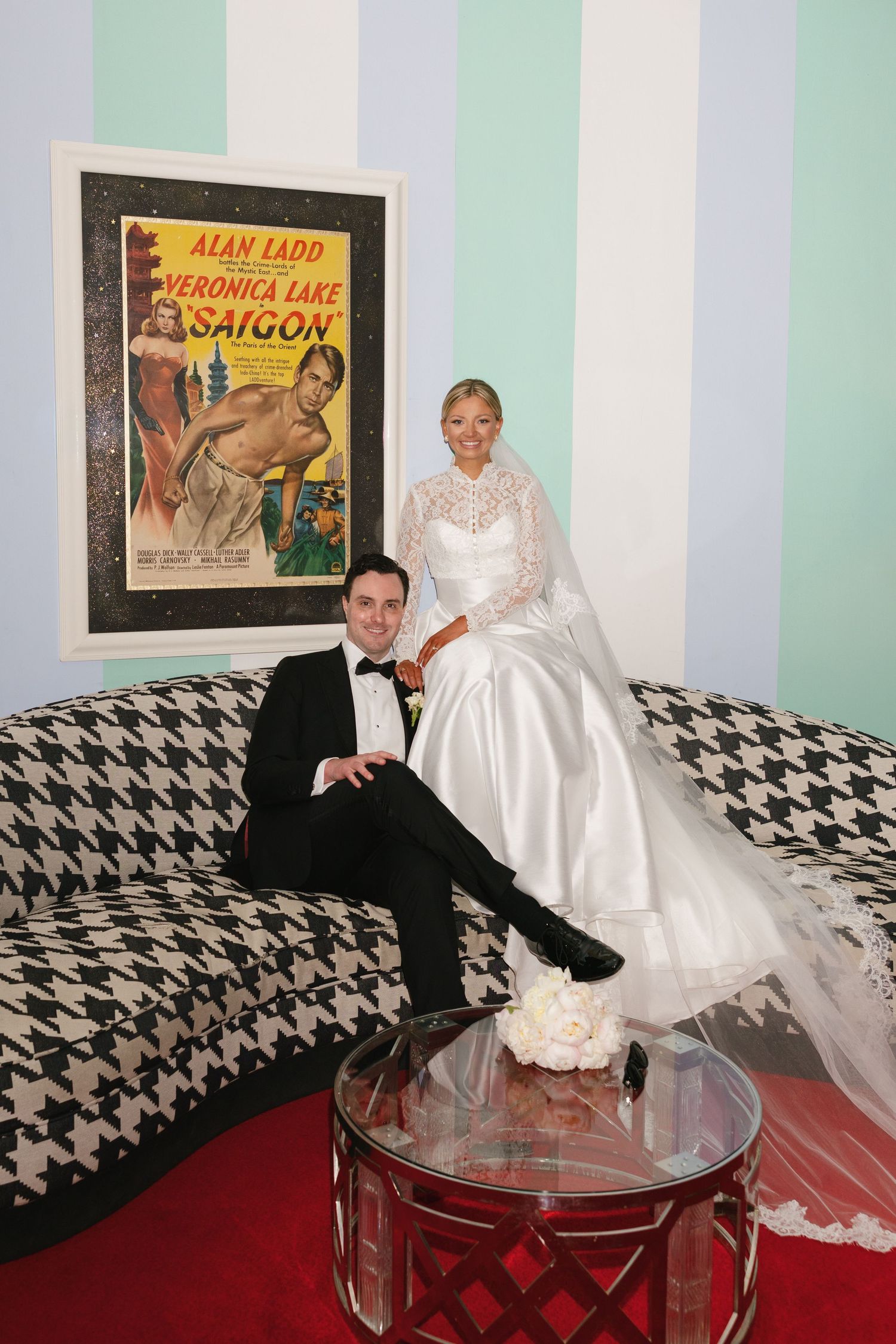 Bride and groom pose elegantly by a vintage Saigon movie poster on a houndstooth sofa at their wedding.