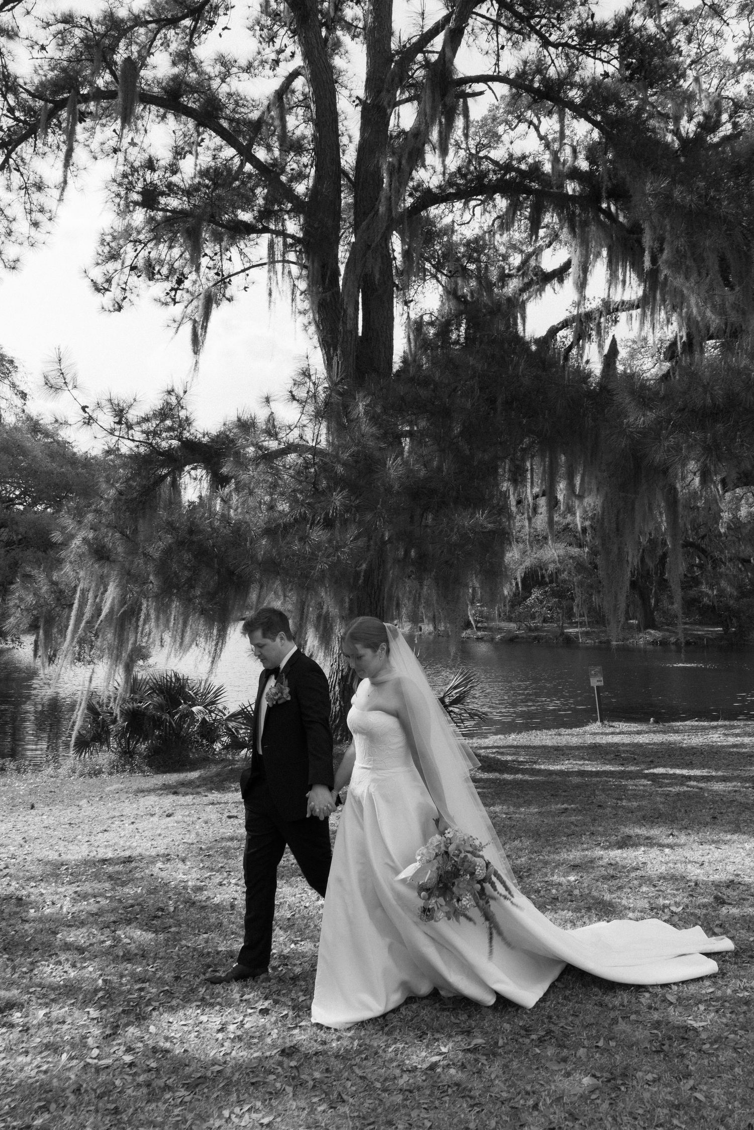 Black and white photo of bride and groom walking hand in hand near a pond under Spanish moss-draped trees.