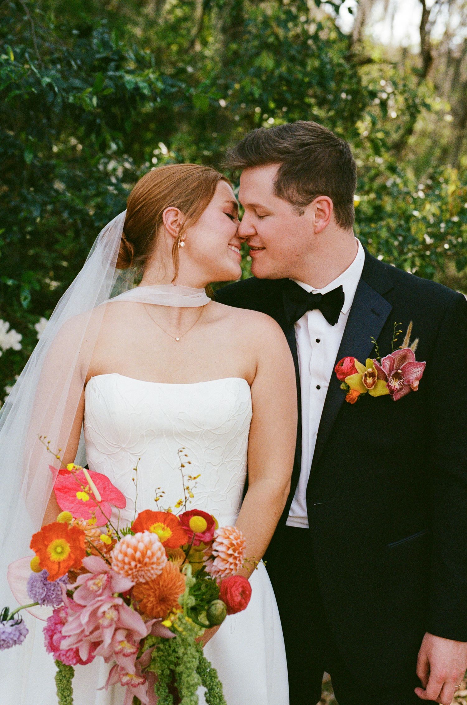 Bride and groom sharing a romantic moment, she holds a vibrant bouquet of orange, pink, and red flowers, he wears a black tuxedo.