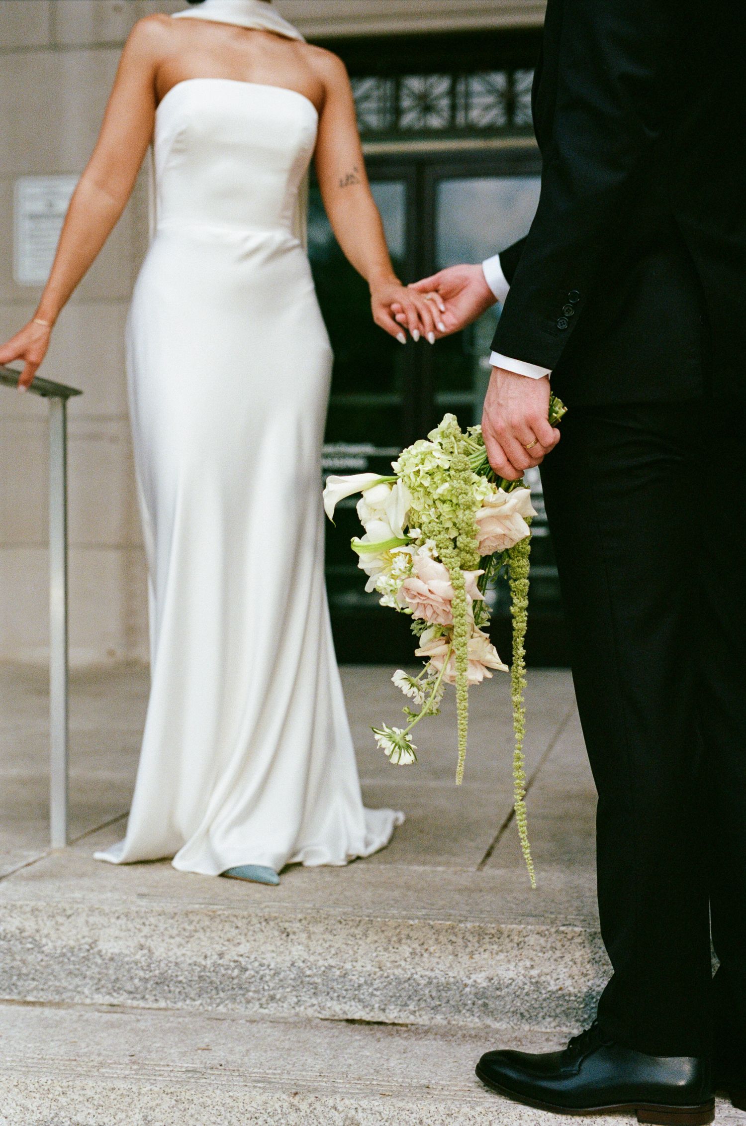 Bride in a strapless white satin gown holds hands with a groom in a black suit, who carries a cascading pink and green bouquet.