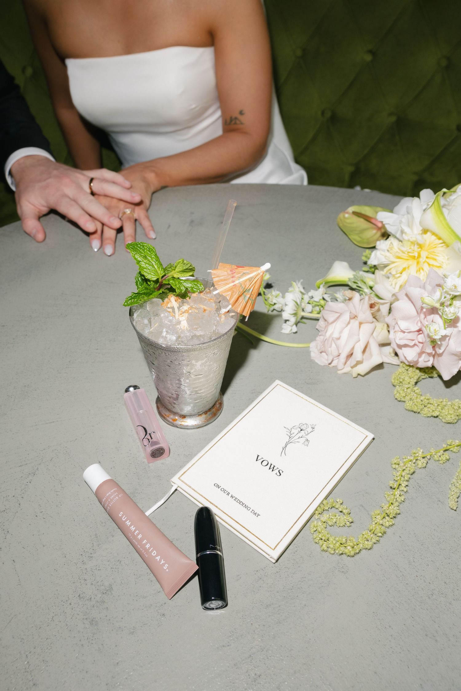 Bride in a strapless white dress holds hands with a partner beside a mint julep, wedding vows booklet, lip products and a pastel bouquet.