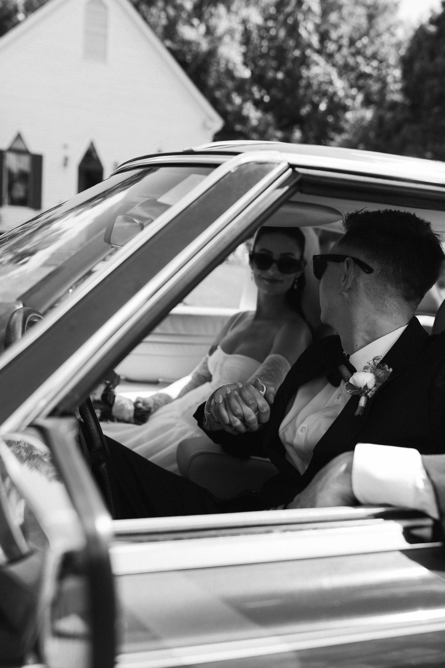 Black and white photo of a bride and groom in sunglasses holding hands inside a vintage car, with a white building and trees behind.