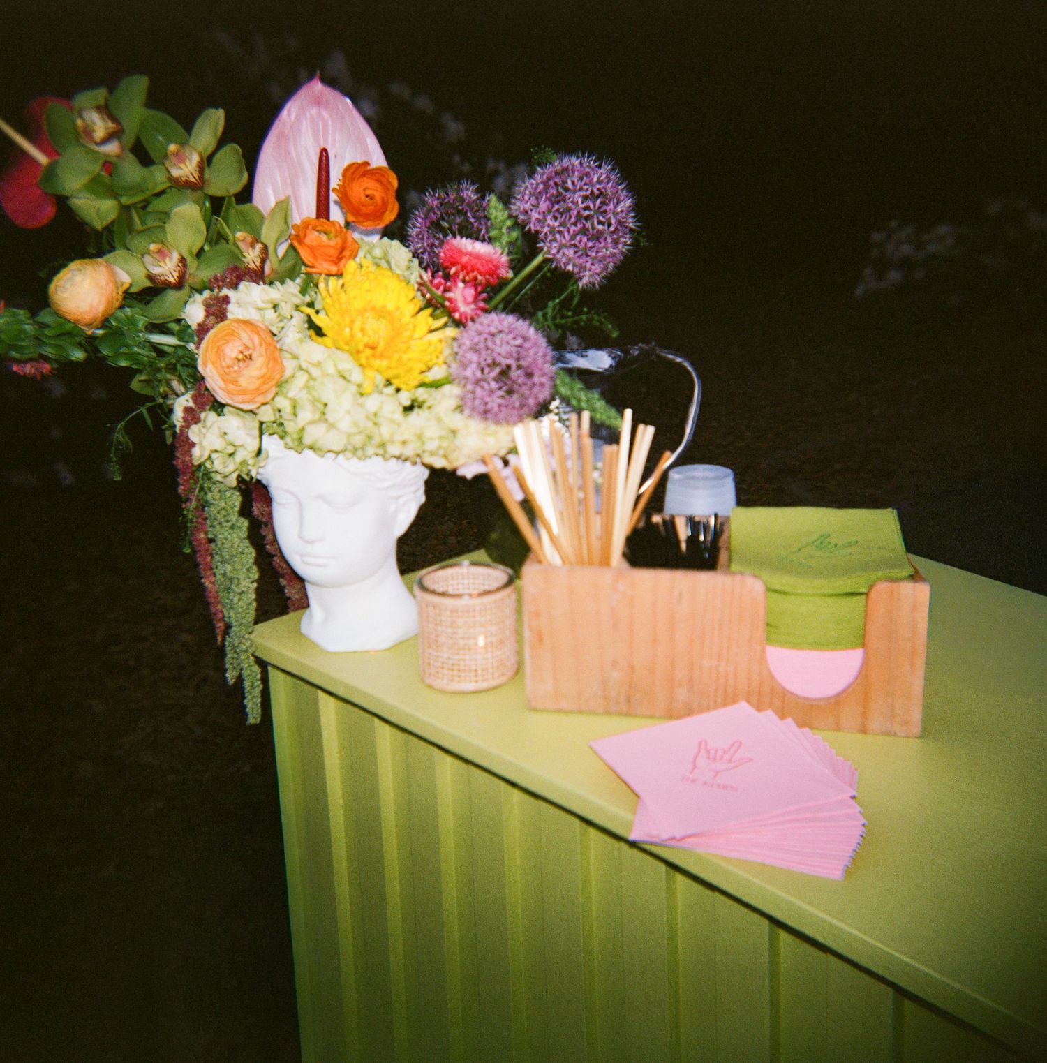 Colorful floral arrangement in white vase on lime green table with wooden organizer and pink cards.