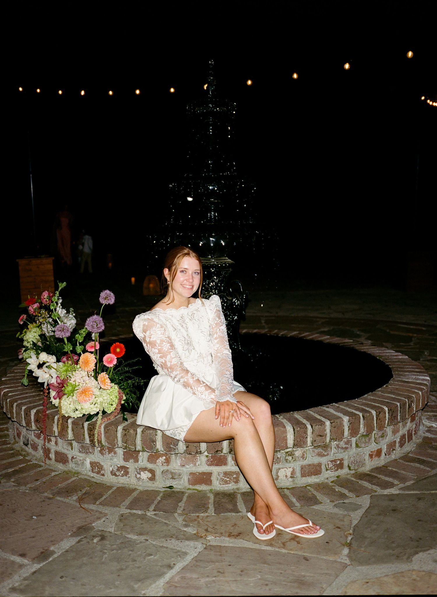 Woman in white lace dress sitting on brick fountain edge at night with colorful floral arrangement and string lights.
