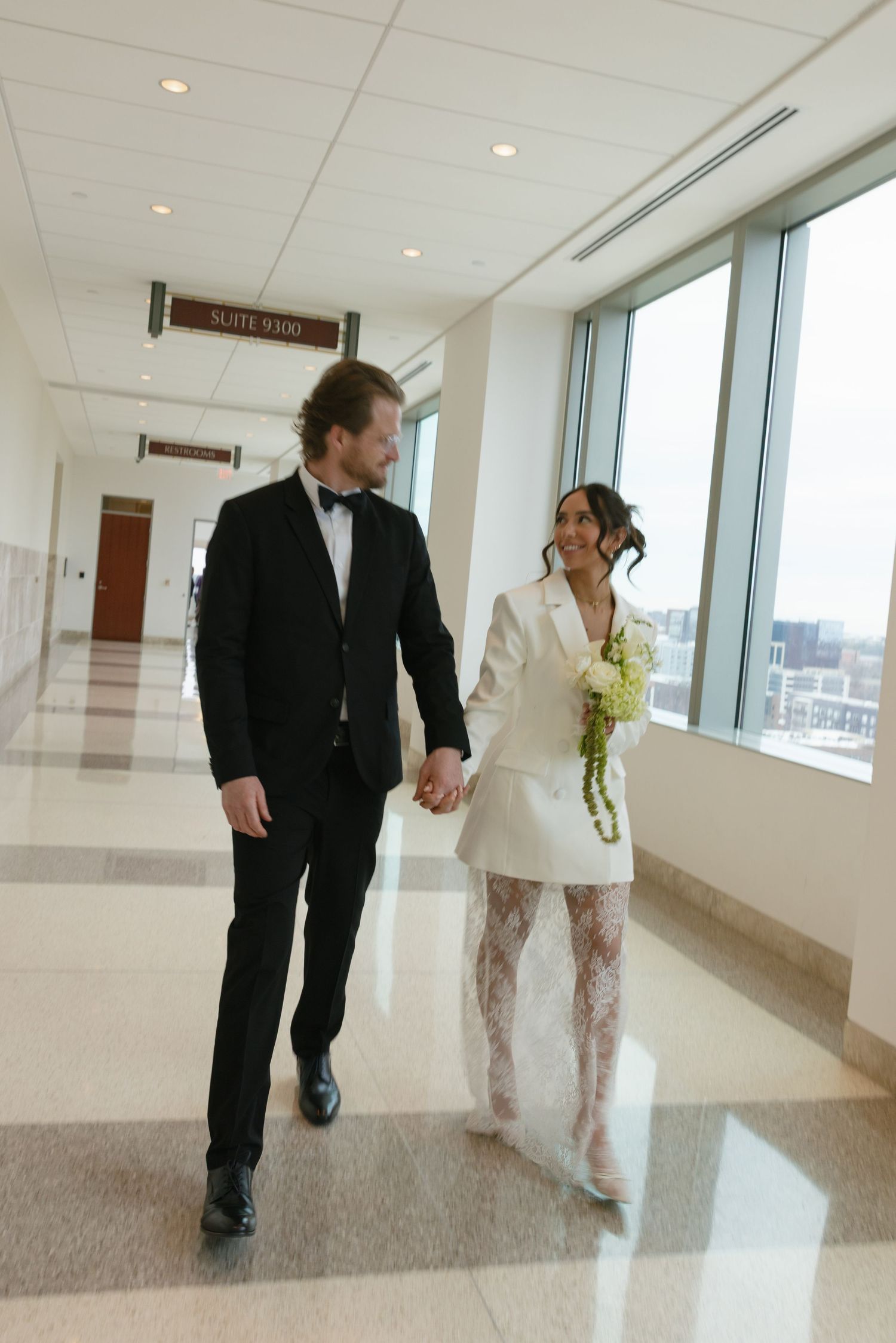 Bride in white blazer dress with lace tights holds bouquet while walking hand-in-hand with groom in black tuxedo.