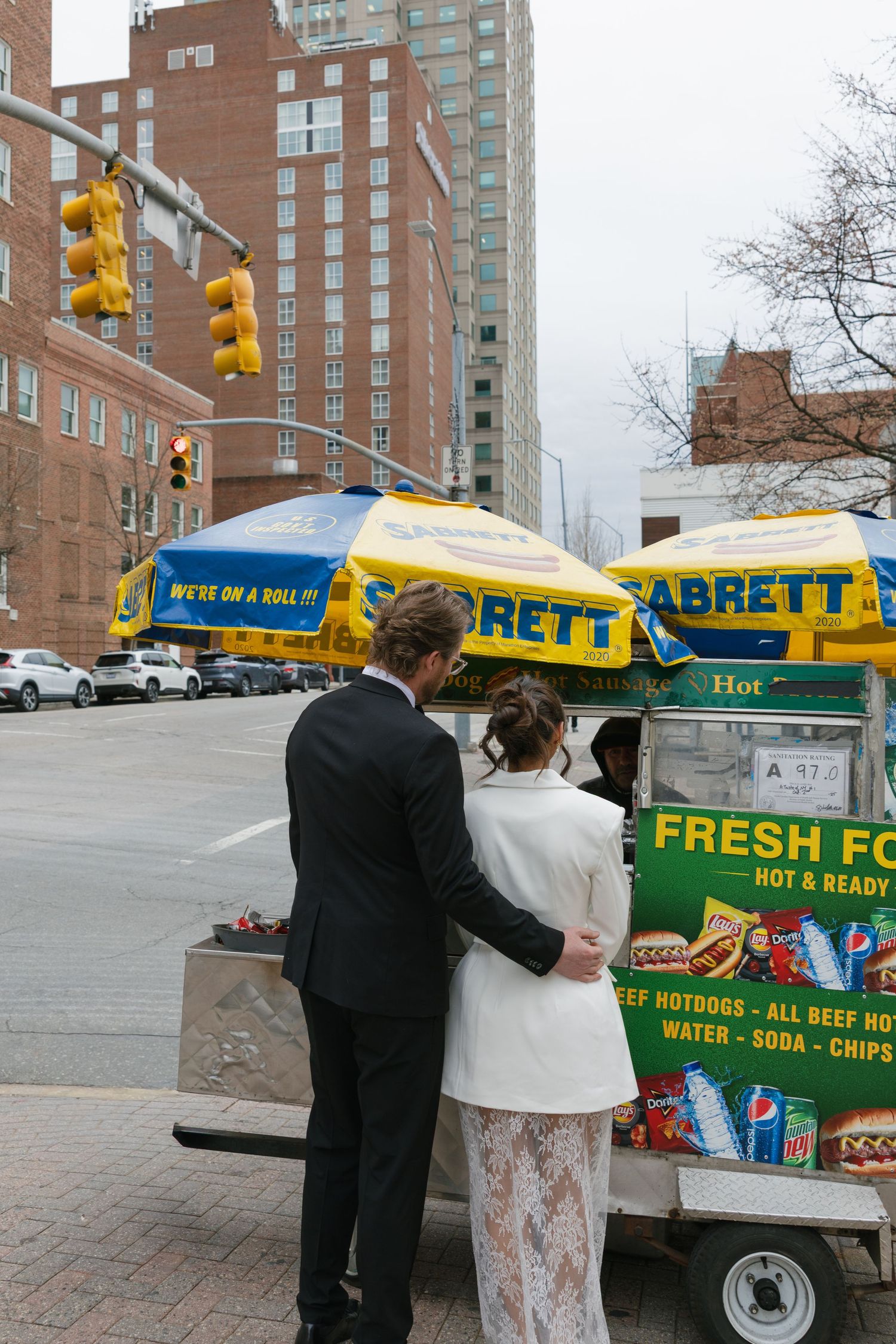 Bride and groom in wedding attire stopping at a Sabrett hot dog cart on a New York City street.