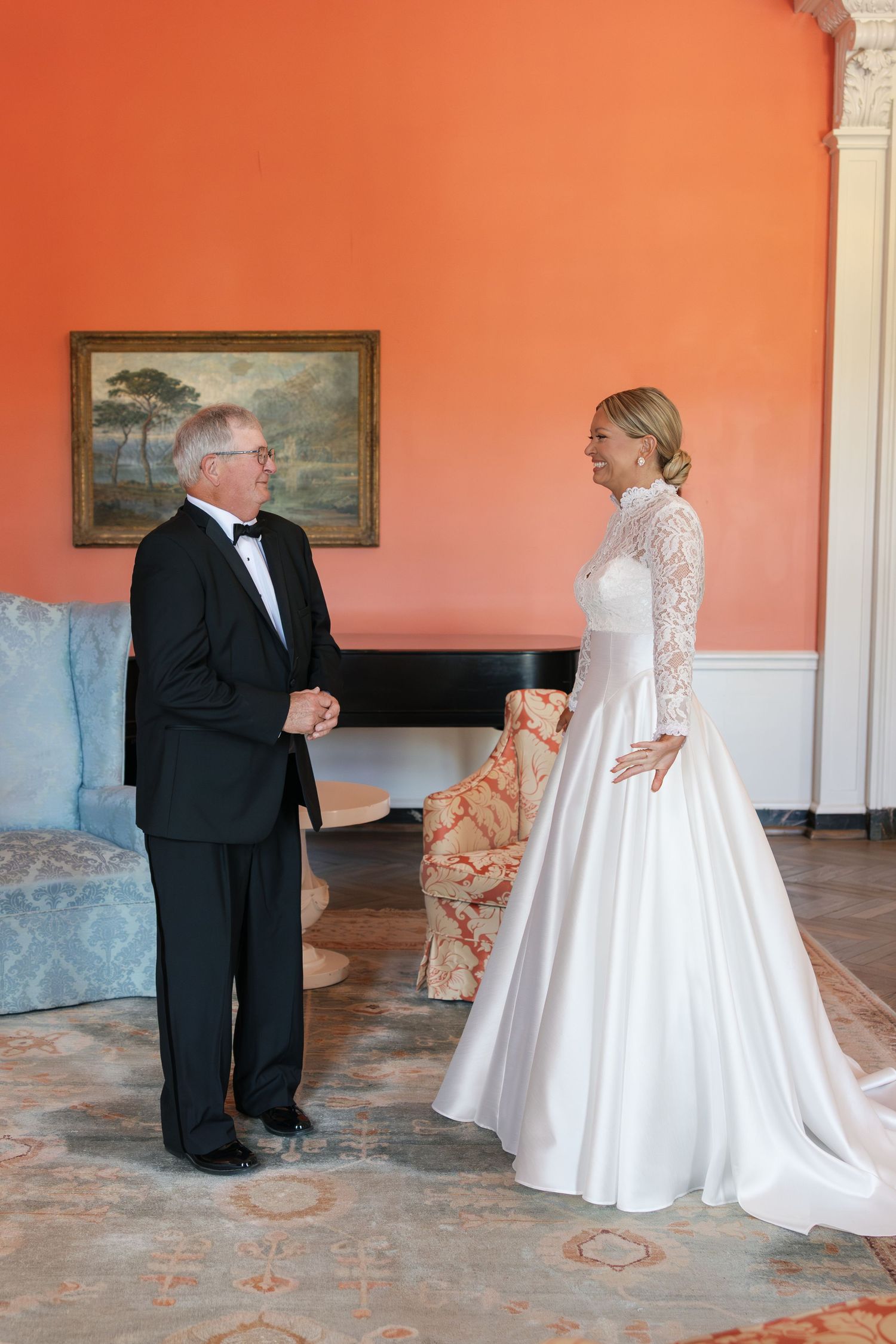 Bride in lace long-sleeve ball gown shares first look moment with father in tuxedo in elegant orange room.