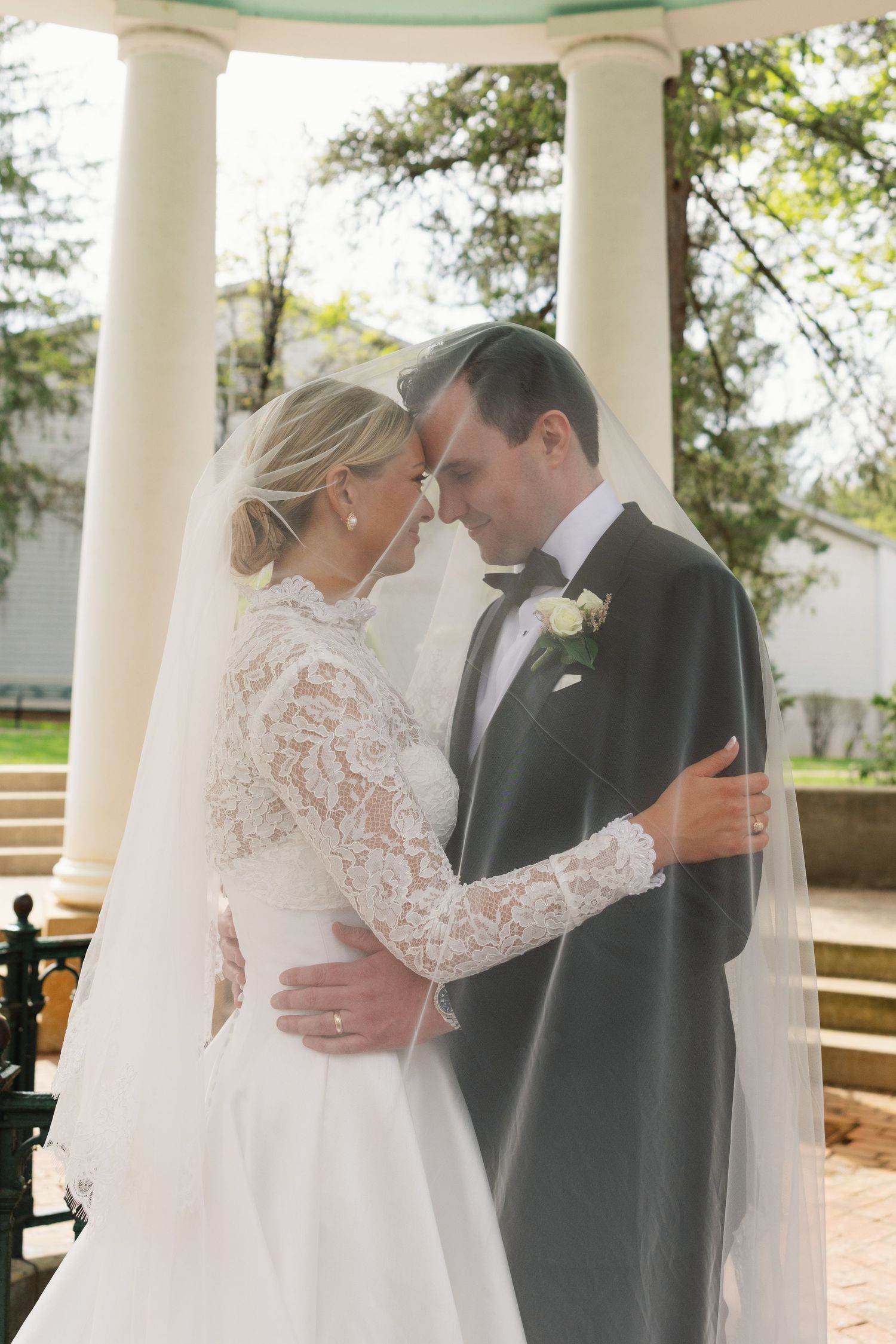 Bride and groom in elegant wedding attire embrace tenderly under a white gazebo surrounded by lush greenery.
