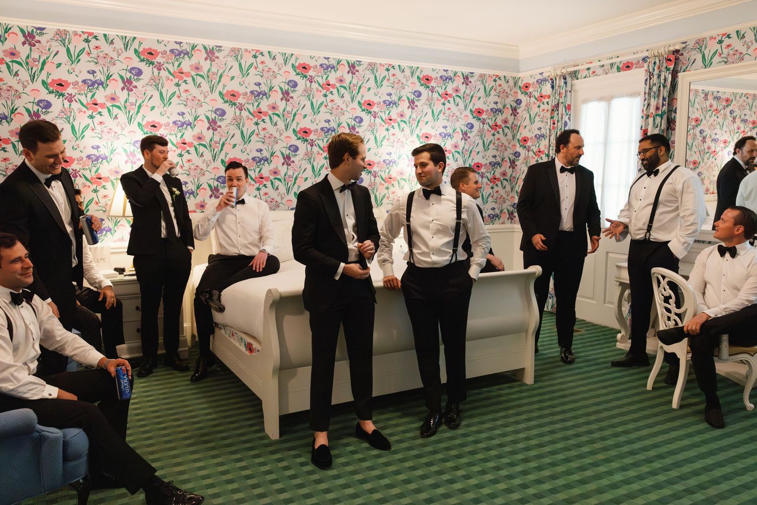 Groomsmen in black tuxedos gather in a floral-wallpapered hotel room before a wedding ceremony.