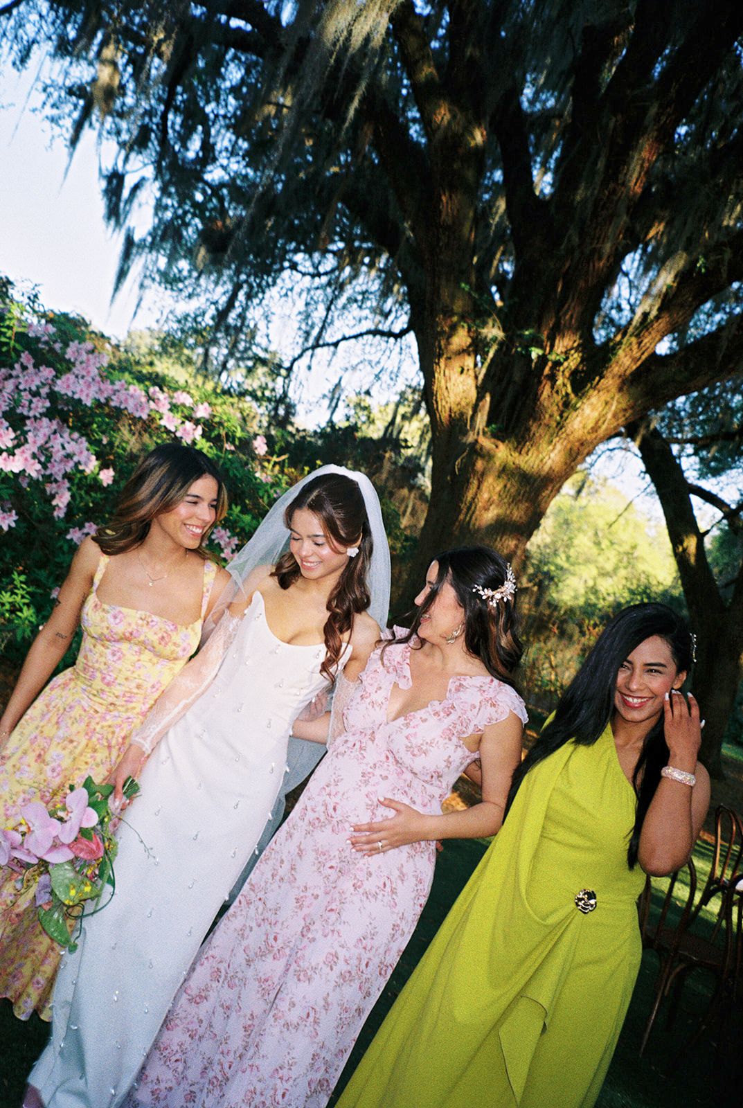 Friends in floral pink and chartreuse dresses share joyful laughs during outdoor photo session under mossy trees.