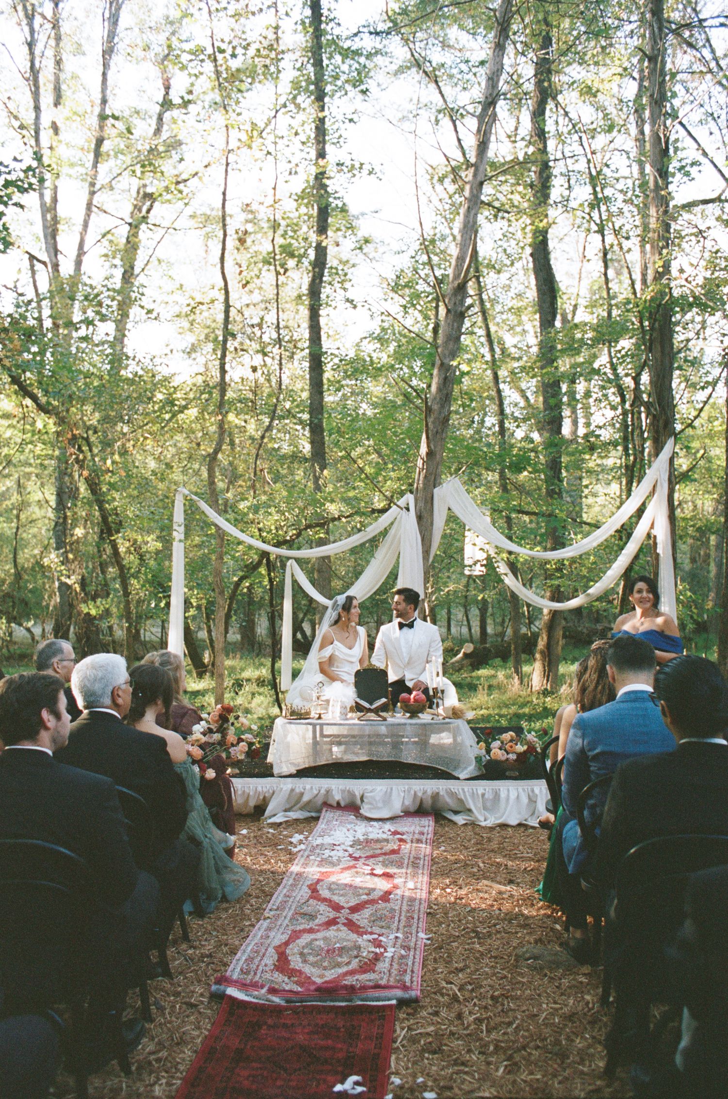 Rustic outdoor wedding ceremony in a wooded setting with white drapes and oriental rugs decorating the aisle.