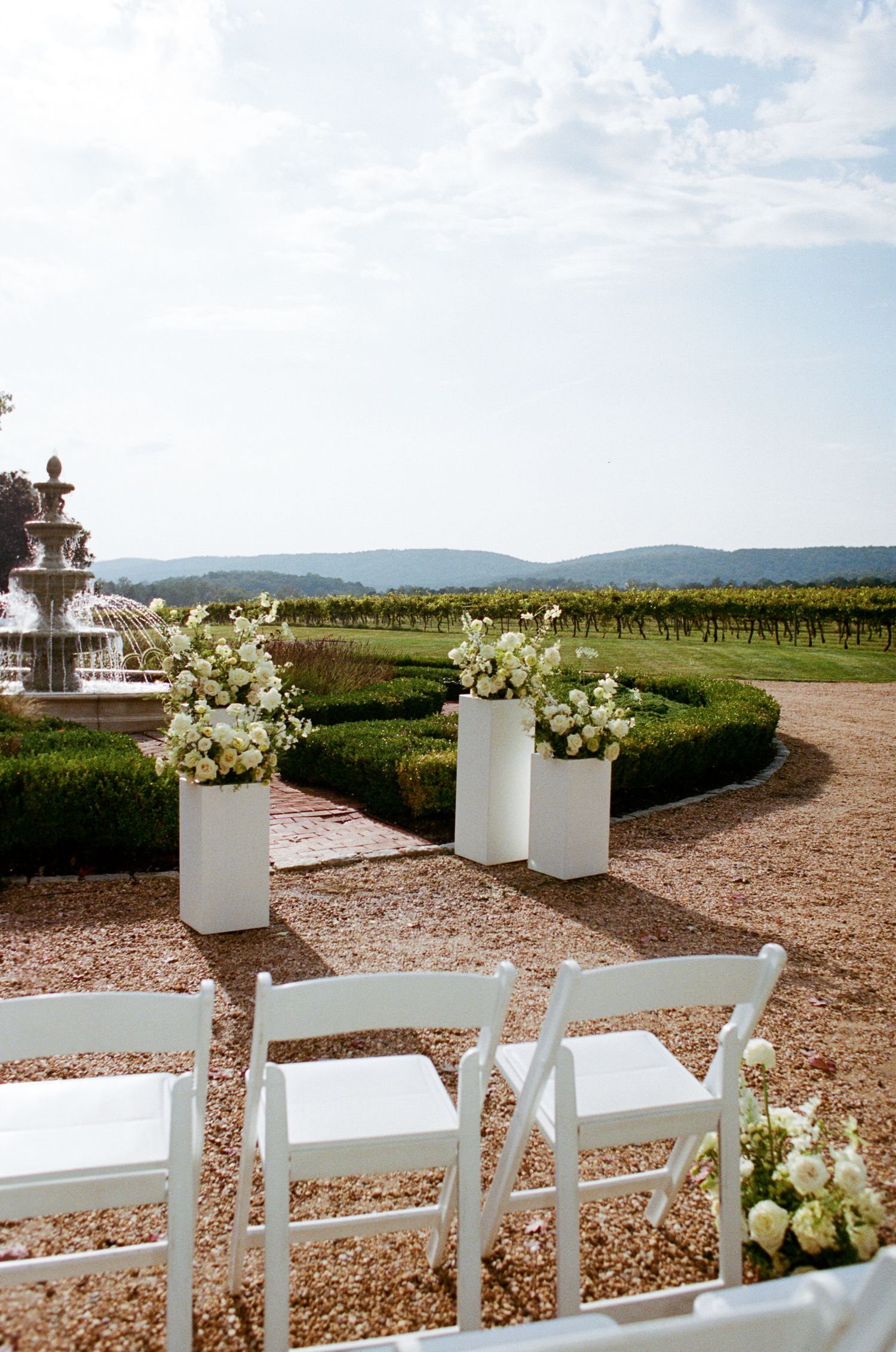 White wedding ceremony setup with floral arrangements and chairs overlooking scenic vineyard and rolling hills.