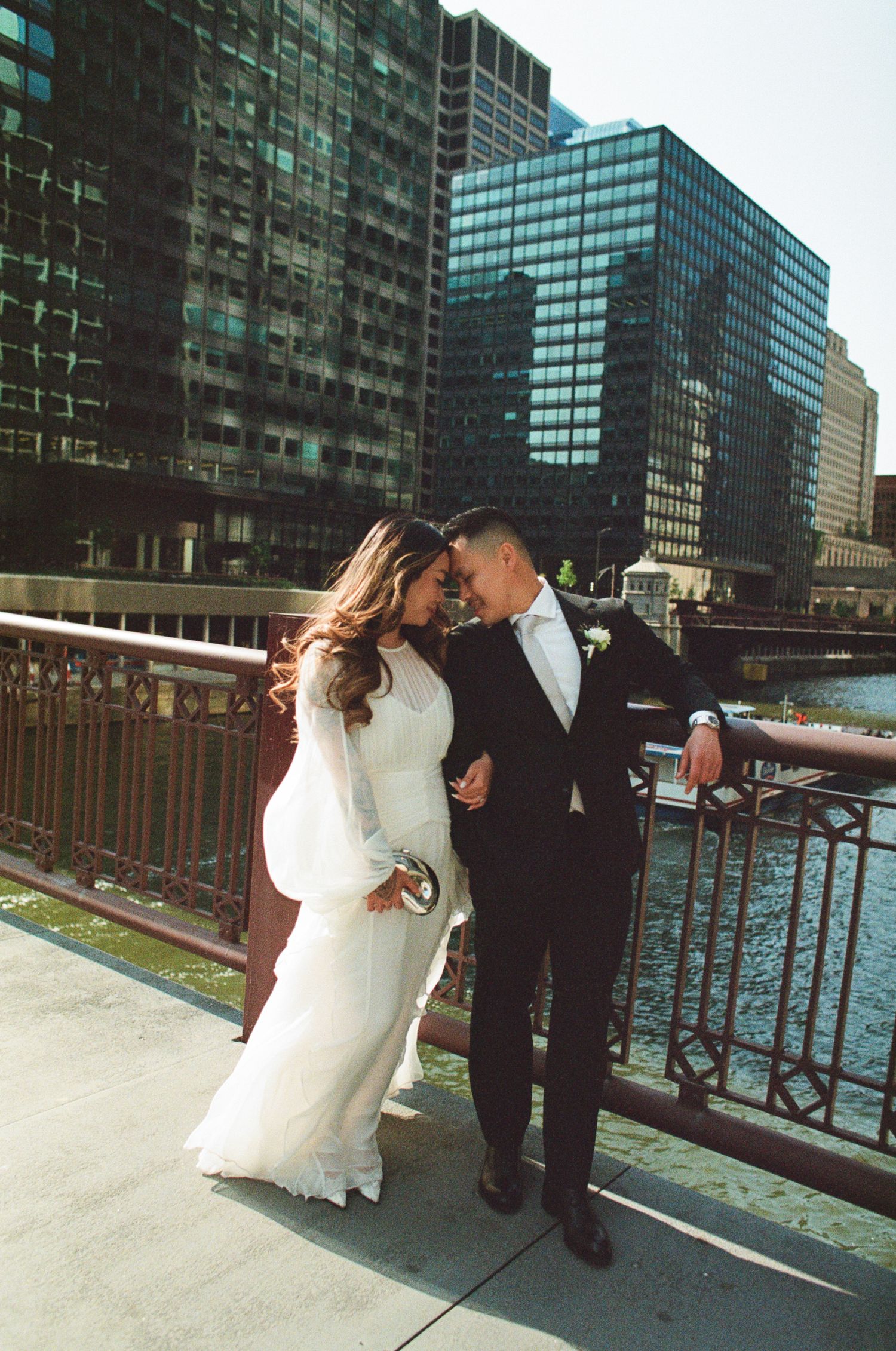A couple shares a romantic moment along a waterfront with modern glass buildings in the background.