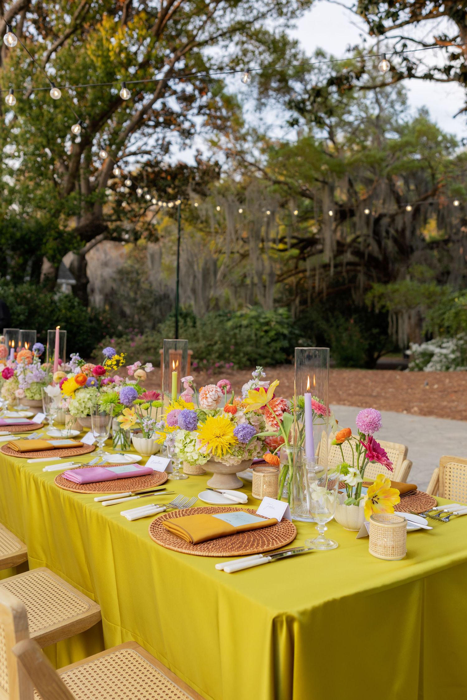 Elegant outdoor dining table with yellow tablecloth, colorful floral arrangements, candles, and wicker chairs under string lights.