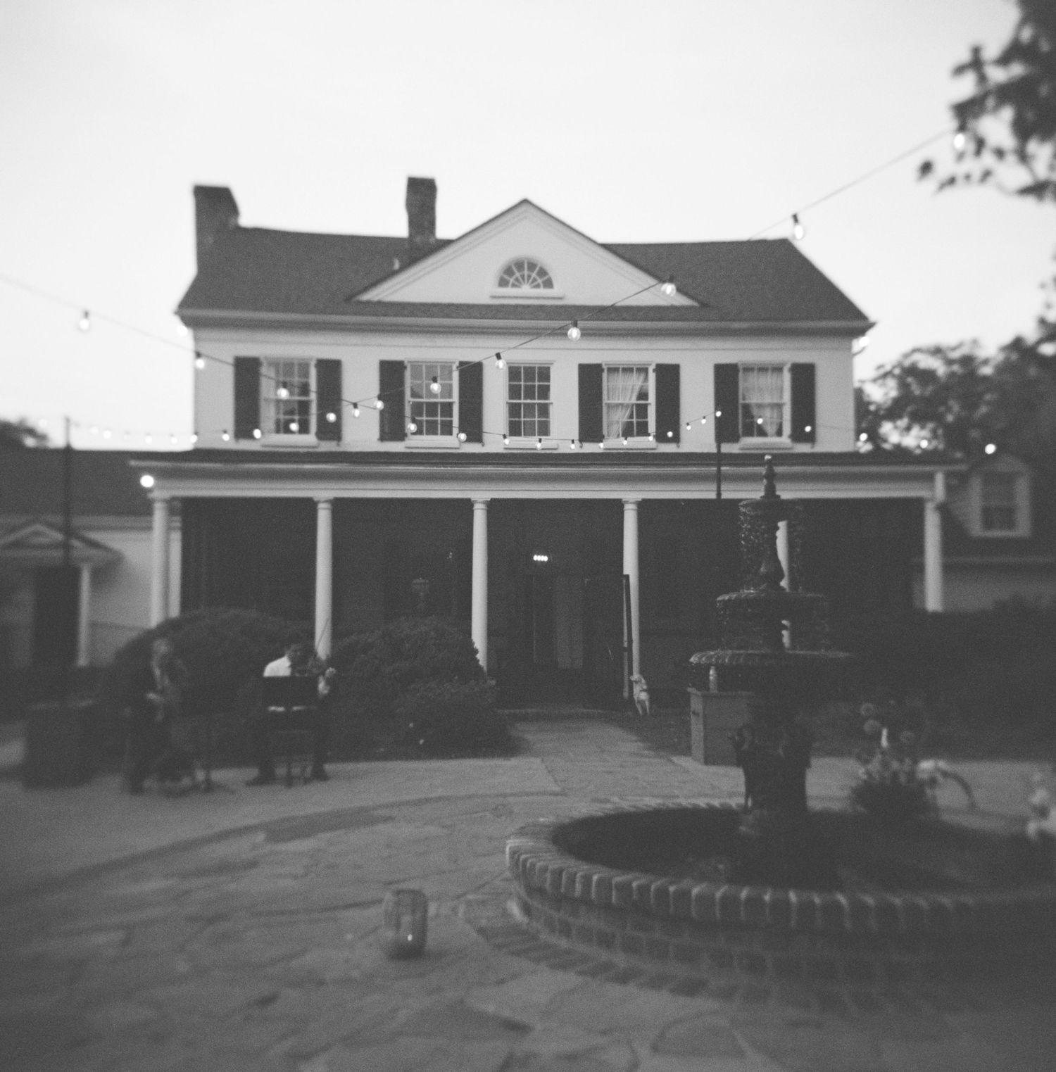 Black and white photo of a colonial-style two-story building with columns and a fountain in the foreground at dusk.