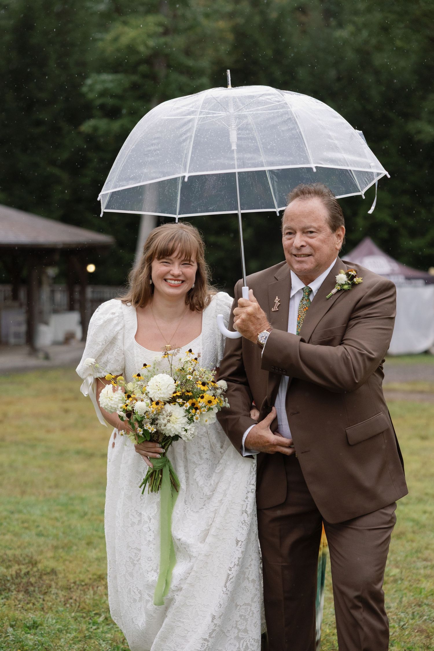 Two people share a clear umbrella while laughing together at an outdoor wedding ceremony in a grassy field.
