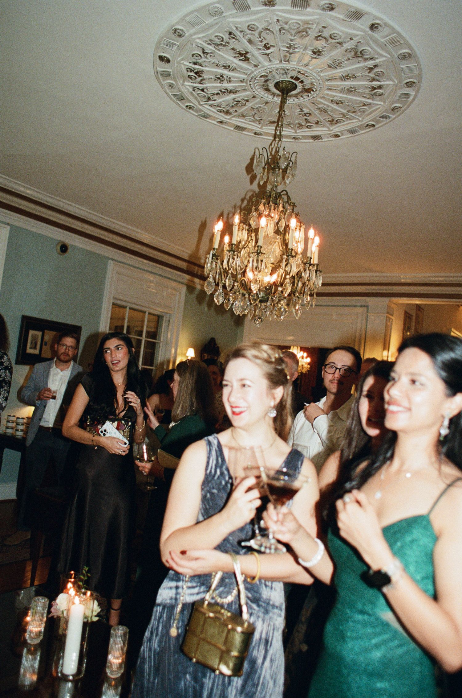 Smiling women in cocktail dresses hold wine glasses at an elegant party beneath a crystal chandelier and ornate ceiling medallion.