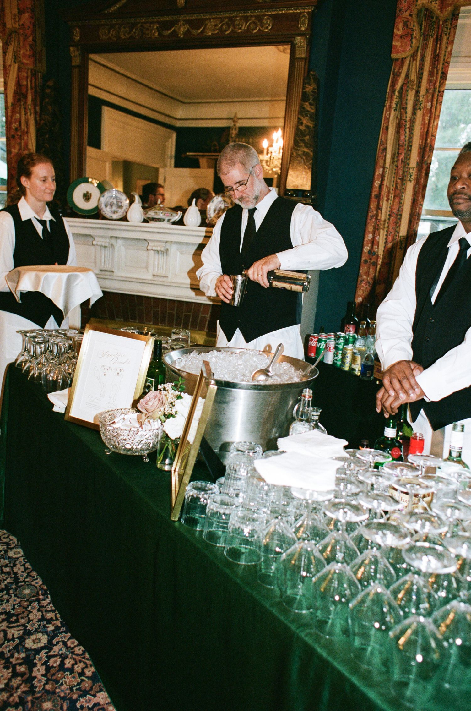 Bartenders in vests and ties pour drinks at an elegant reception bar with rows of glasses, an ice bucket and flowers in a formal ballroom.