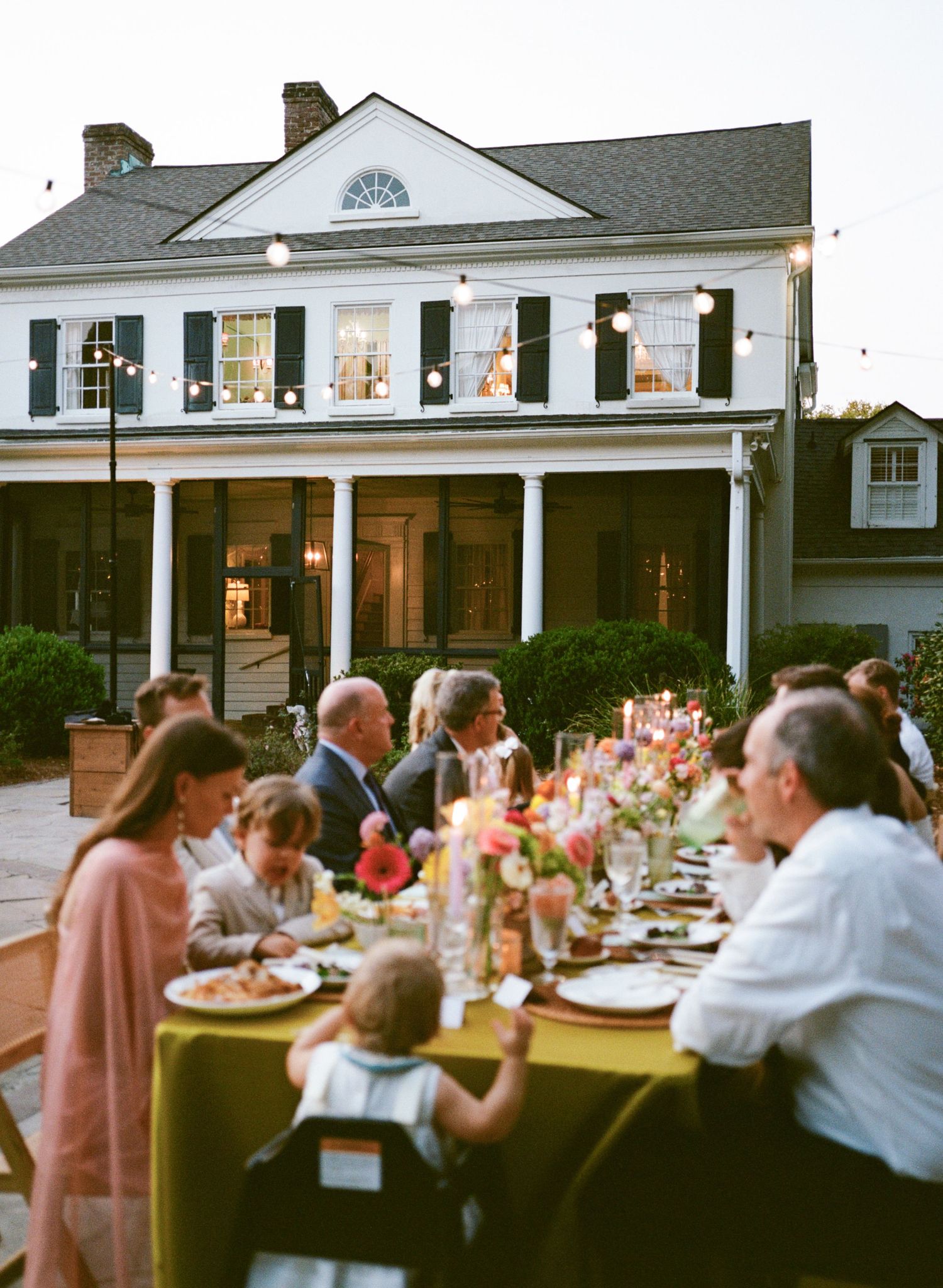 Elegant outdoor dinner party at dusk beside a white colonial home with string lights and guests gathered around a candlelit table.