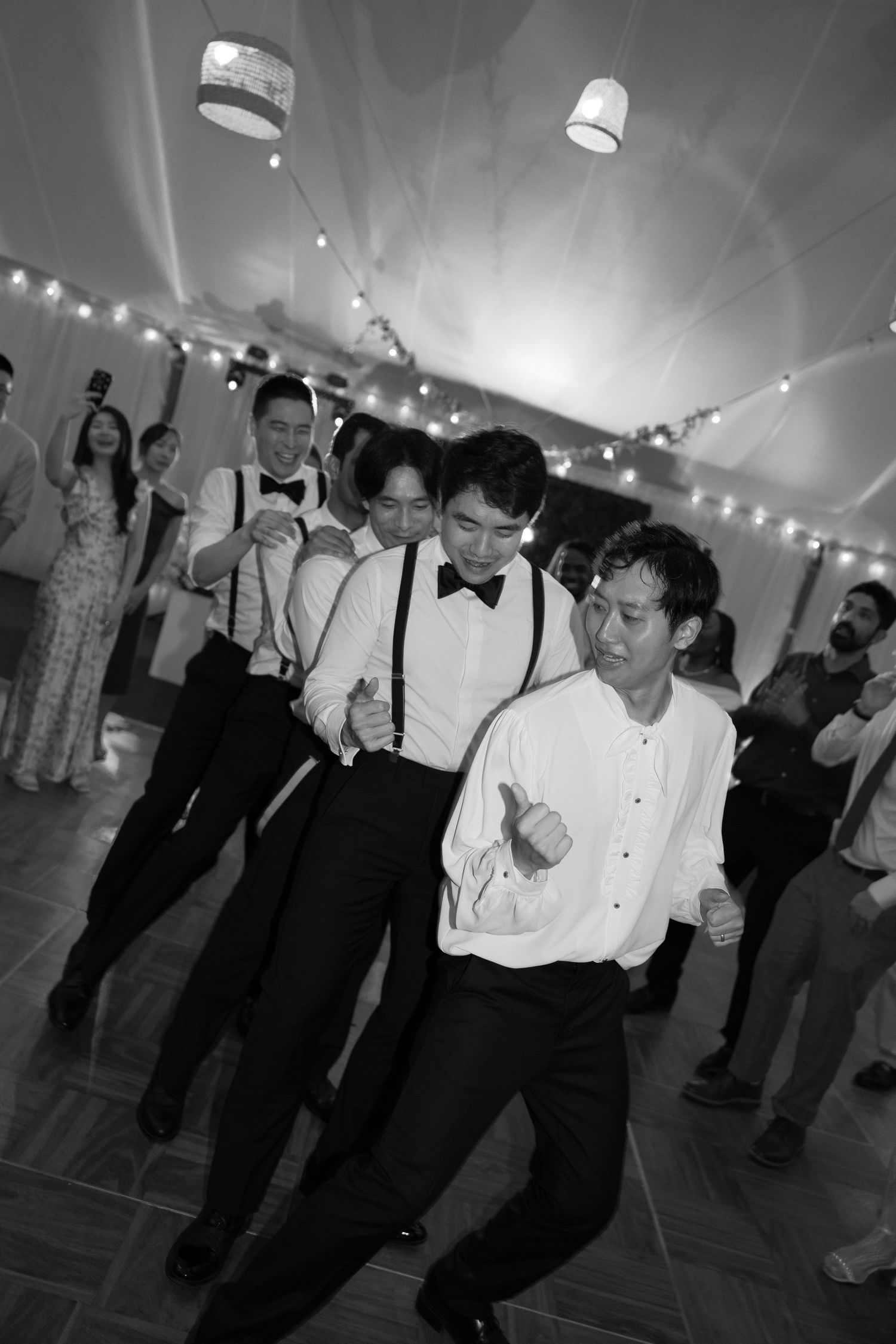 Black and white photo of people dancing at a formal event with string lights overhead and white lamp shades visible.