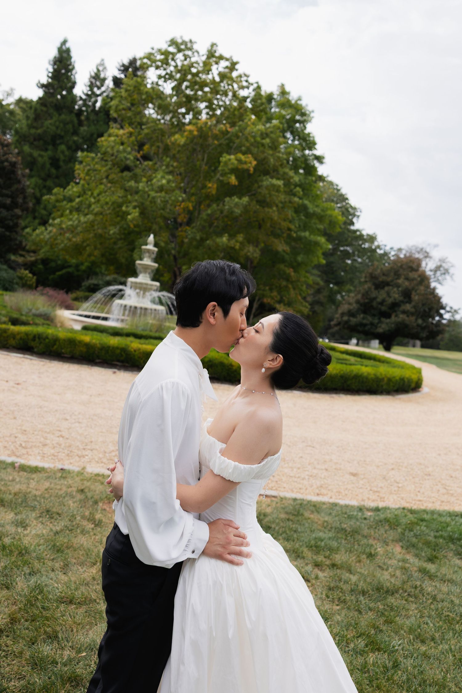 A romantic couple in wedding attire dances and embraces on a grassy lawn with trees in the background.