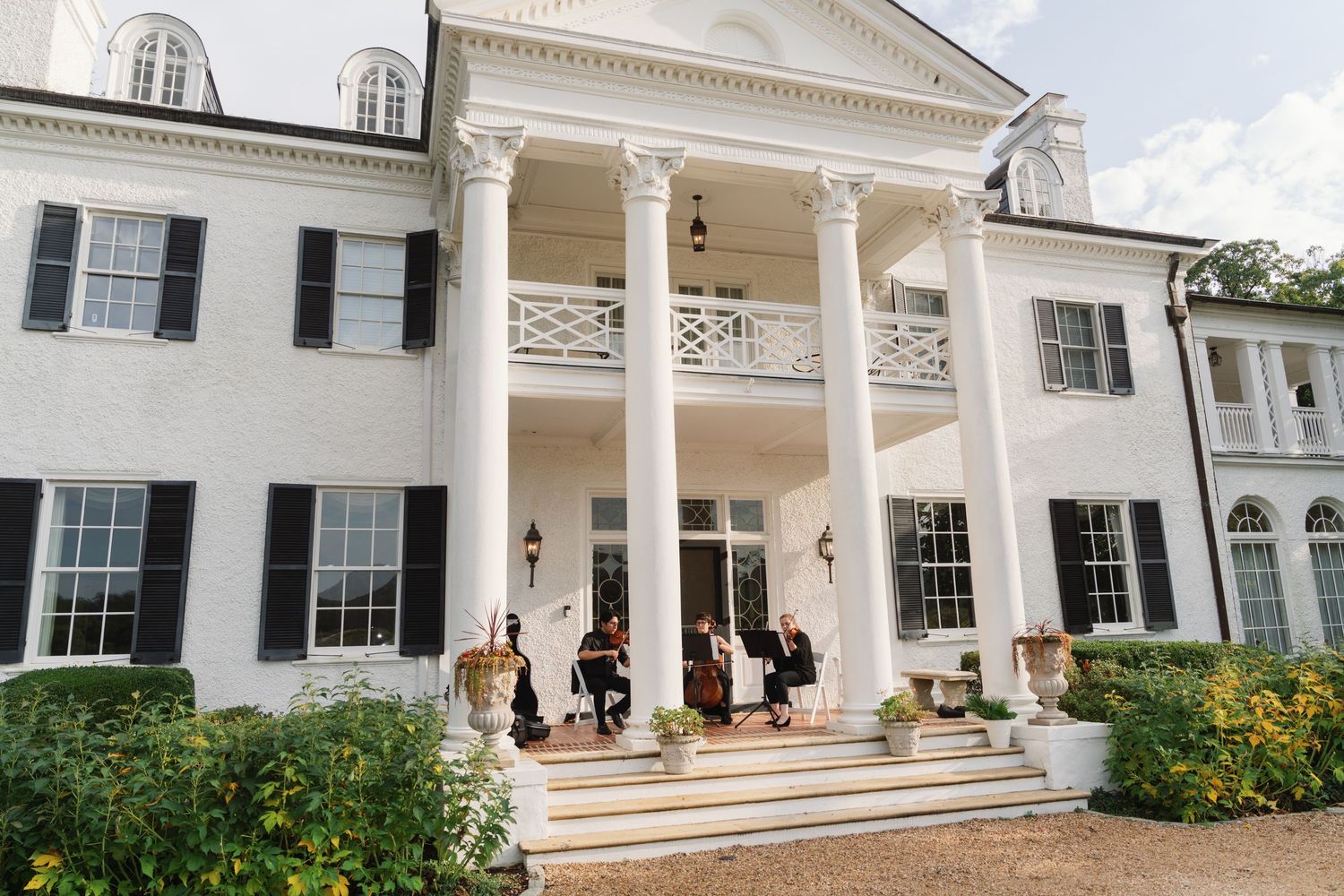 Elegant southern mansion with tall white columns and two-story porch features guests gathering on the front steps.
