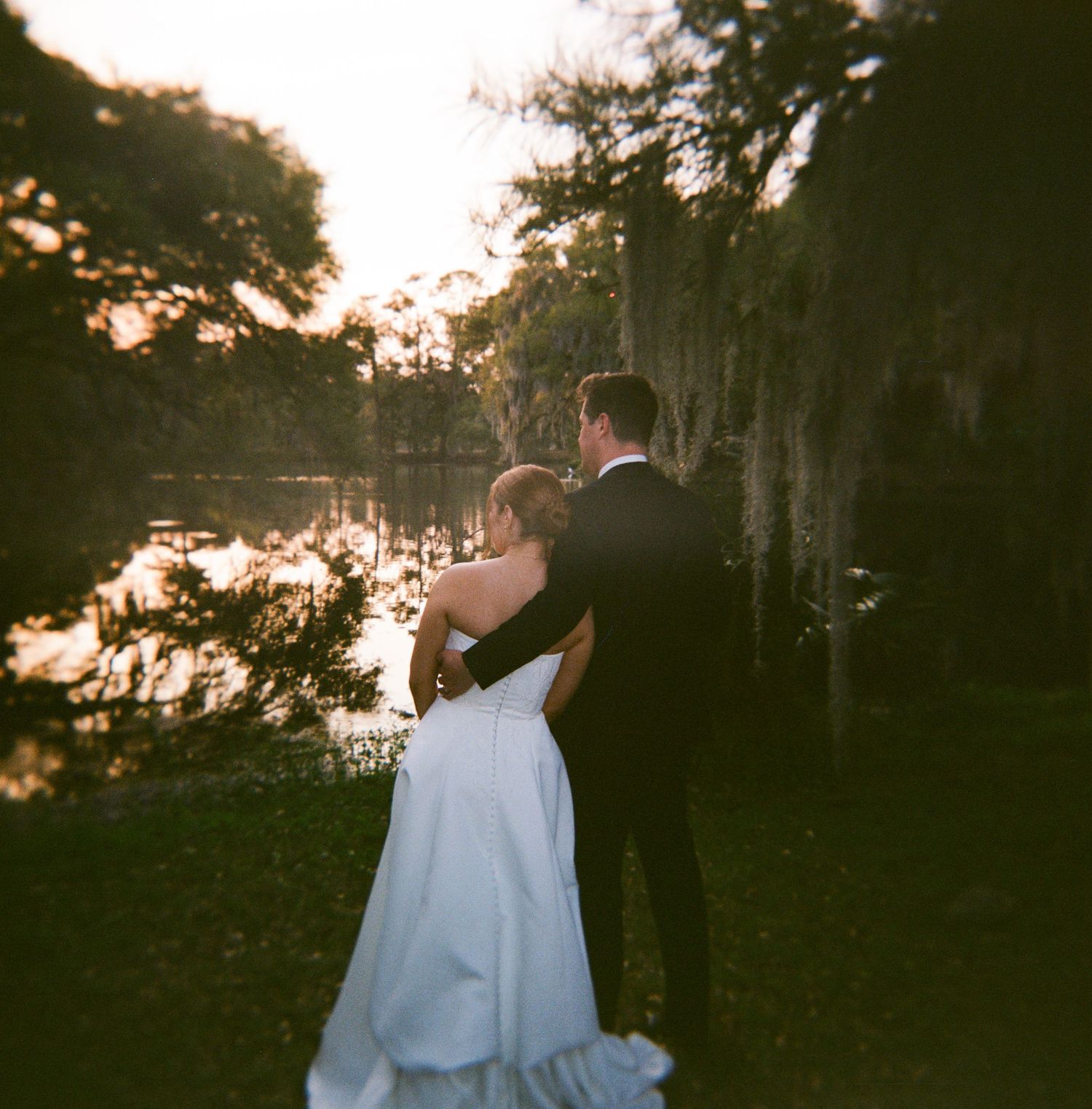A bride and groom stand together gazing at a sunlit pond surrounded by moss-draped trees at their outdoor wedding.