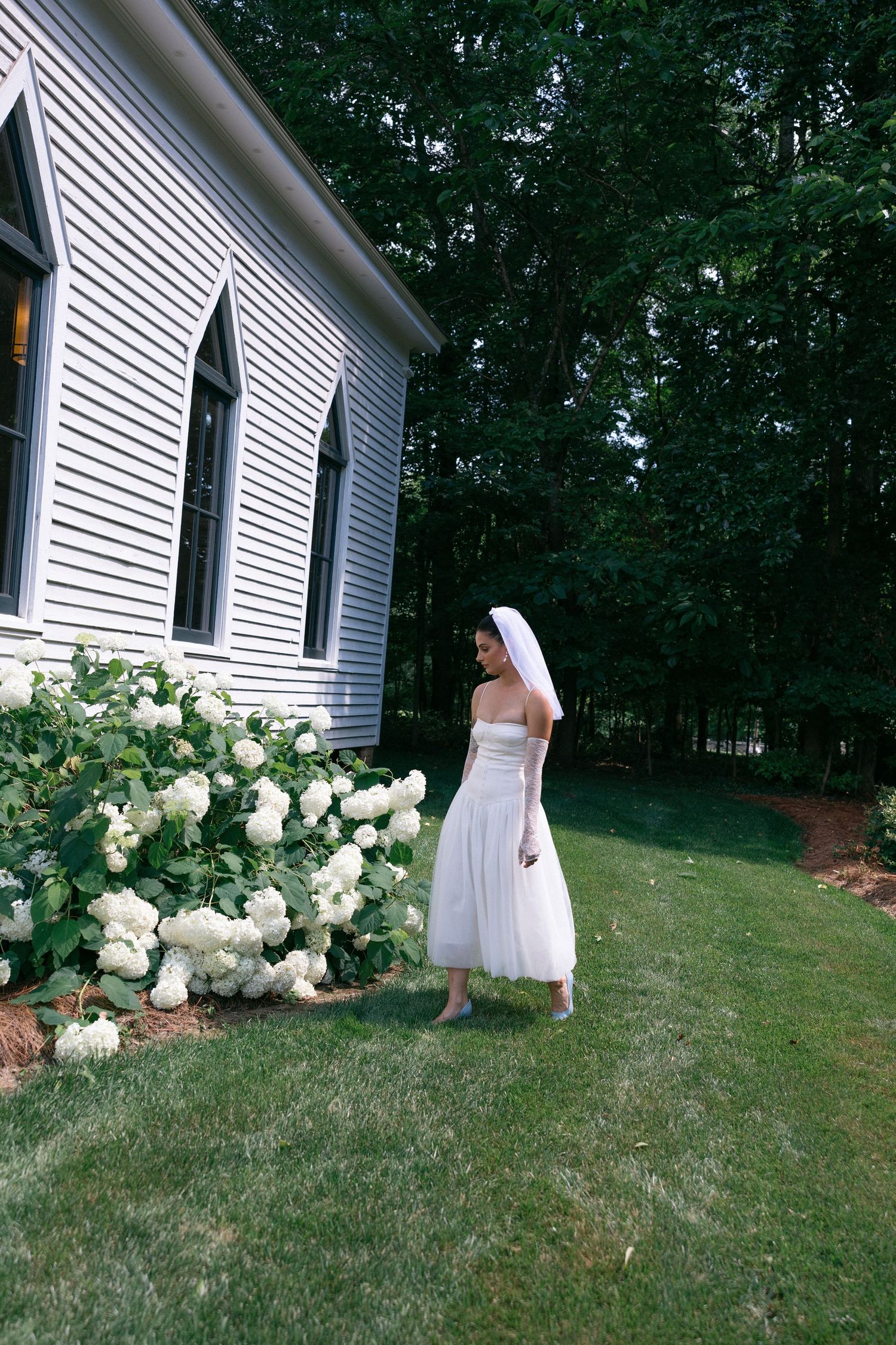 Bride in a white strapless gown and short veil walks across a green lawn beside a white chapel and blooming white hydrangeas.