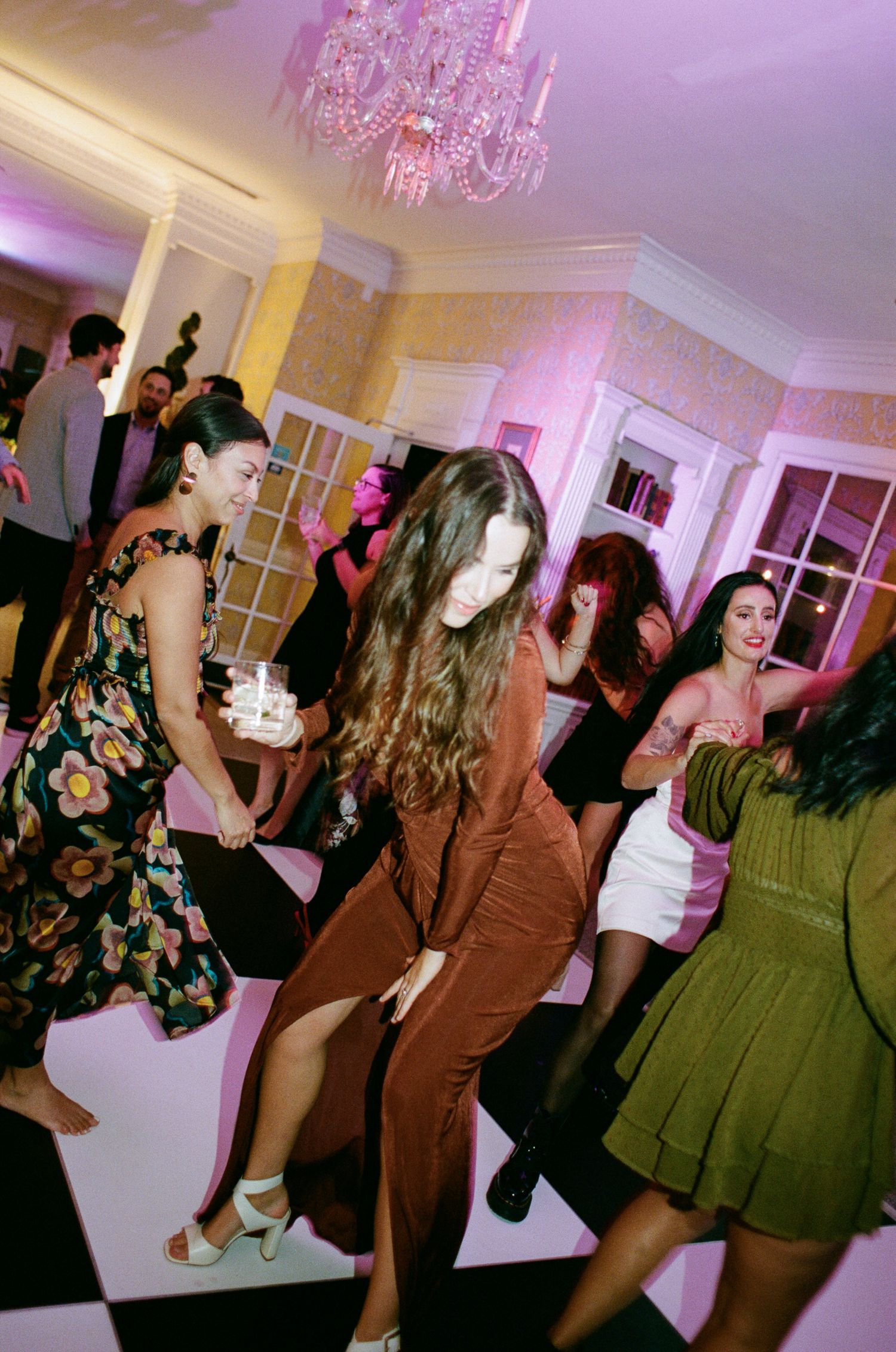 Guests in colorful dresses dance on a black and white checkered floor beneath a crystal chandelier at an elegant indoor party.