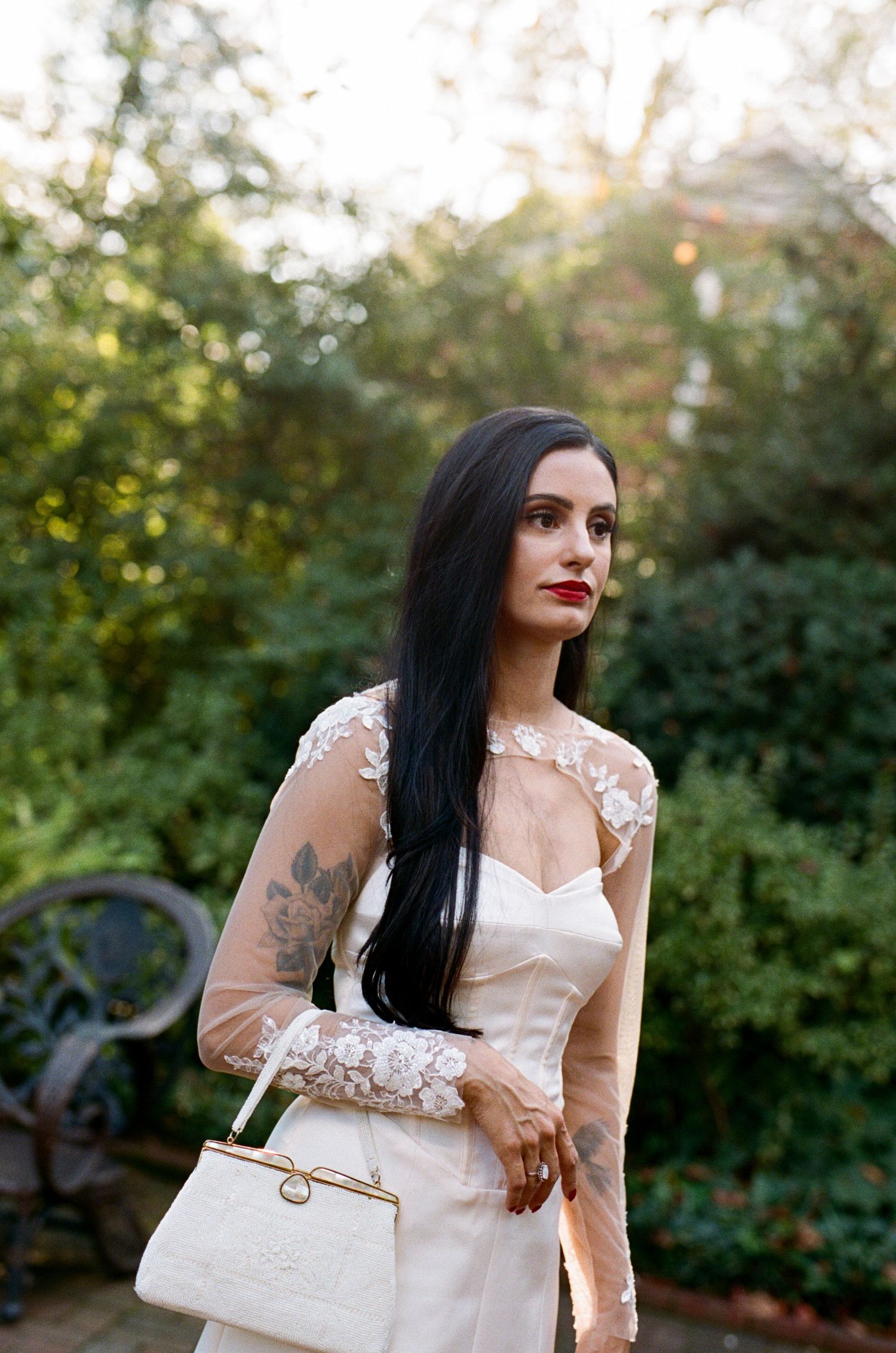 Woman with long dark hair and red lipstick wears a white lace-sleeved bridal gown and holds a vintage white handbag in a lush garden.